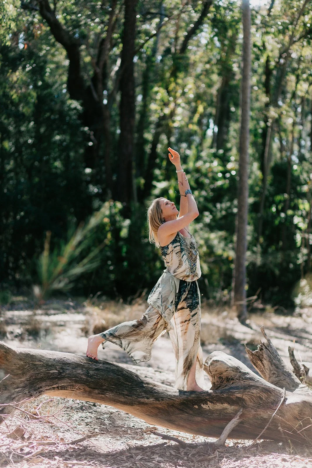 A woman standing on a fallen tree trunk in a forest, reaching upward with one arm, wearing a flowing patterned dress.
