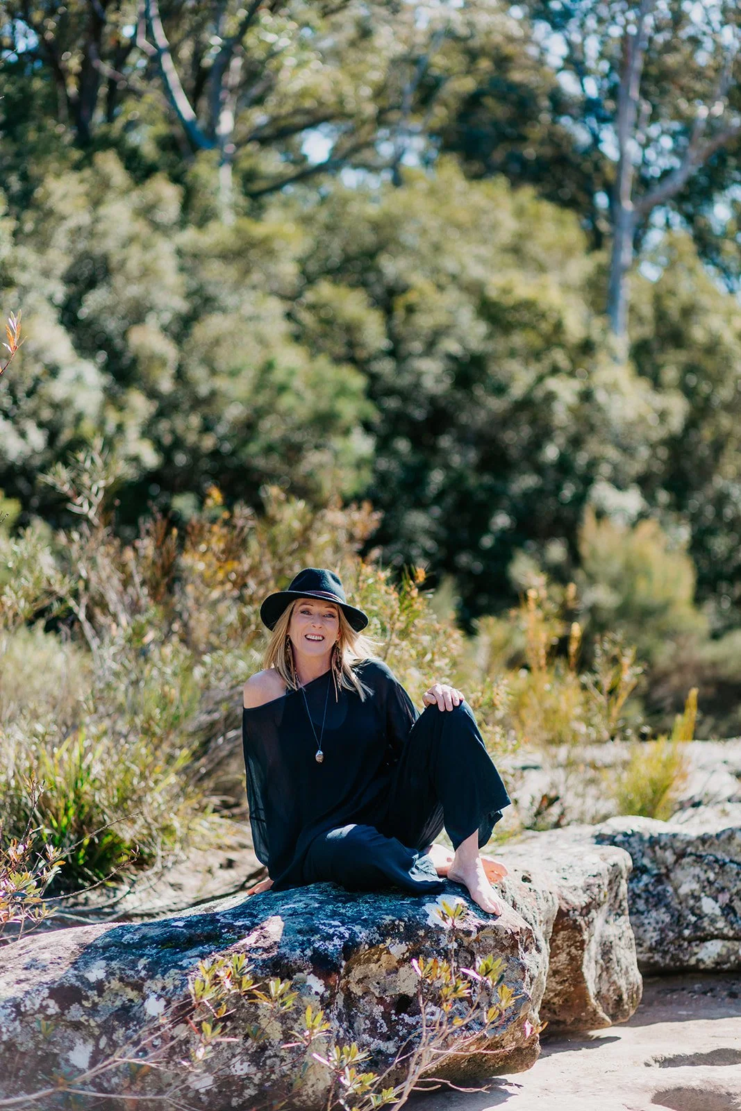 A woman wearing a black dress and wide-brimmed hat sitting on a large rock outdoors, surrounded by greenery and trees, smiling at the camera.
