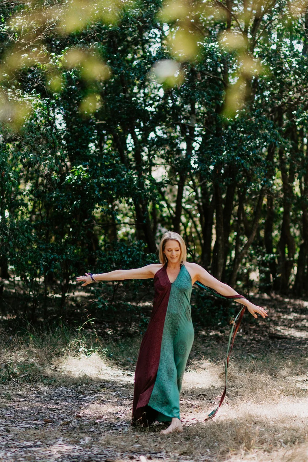 A woman in a colorful, flowing dress dances barefoot in a forest clearing, with her arms extended and a serene expression.