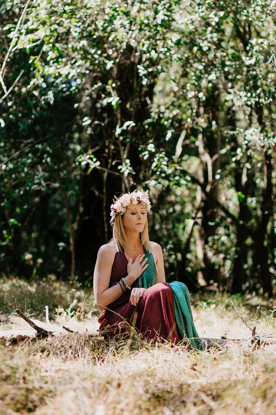 A woman wearing a flower crown, sitting cross-legged outdoors with her eyes closed and hand on her chest, surrounded by greenery and trees in a forest setting.