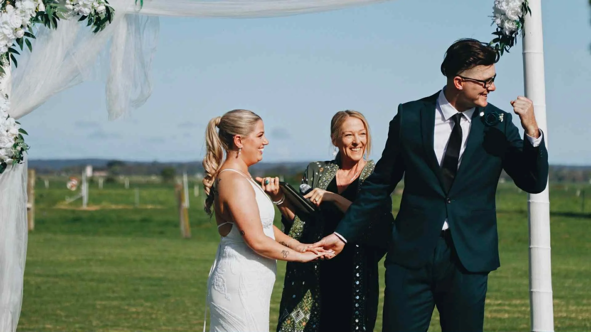 A couple getting married outdoors with an officiant, surrounded by a floral arch, on a sunny day in a grassy field.