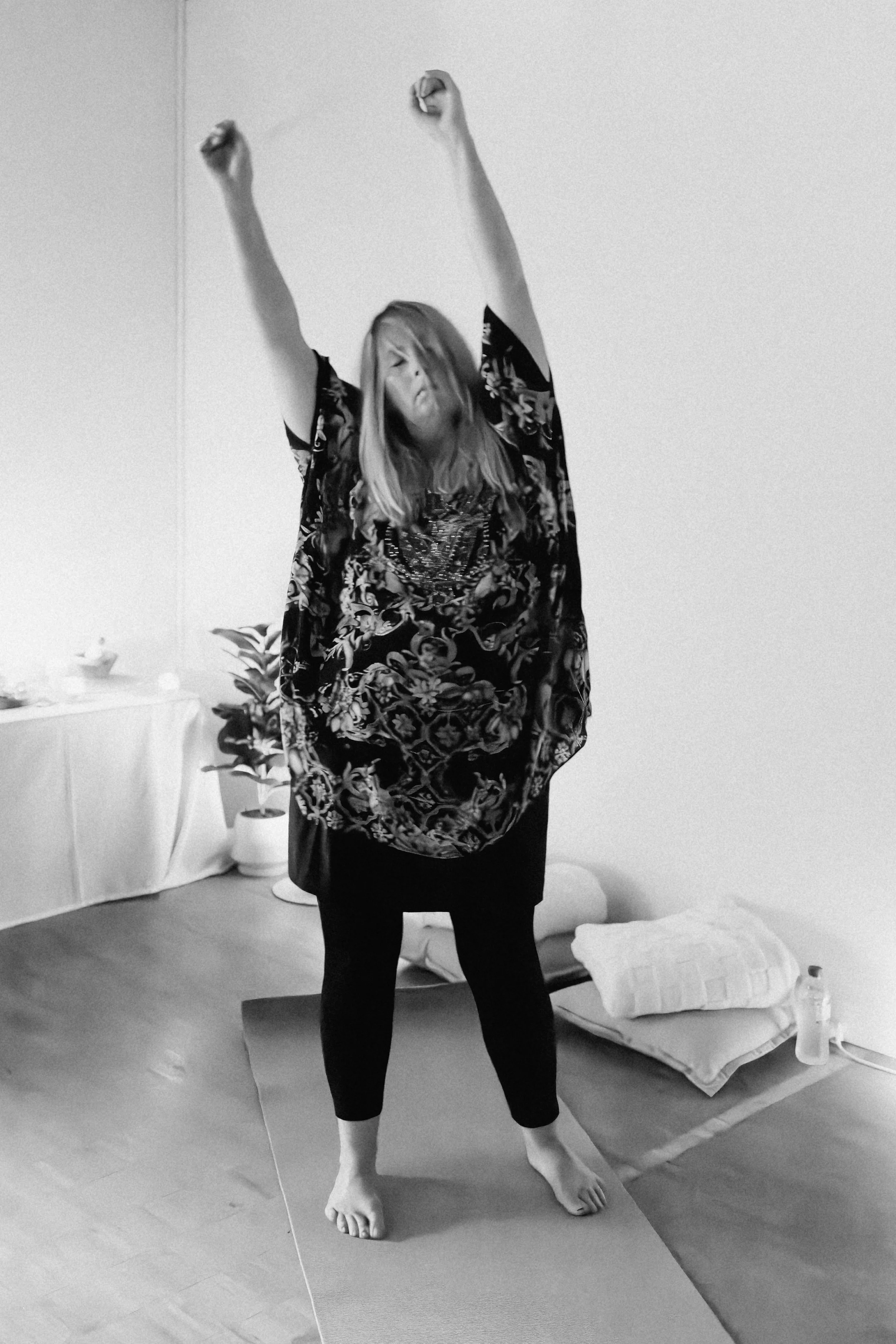A woman standing on a yoga mat stretching her arms above her head in a room with a bed and plants in the background.