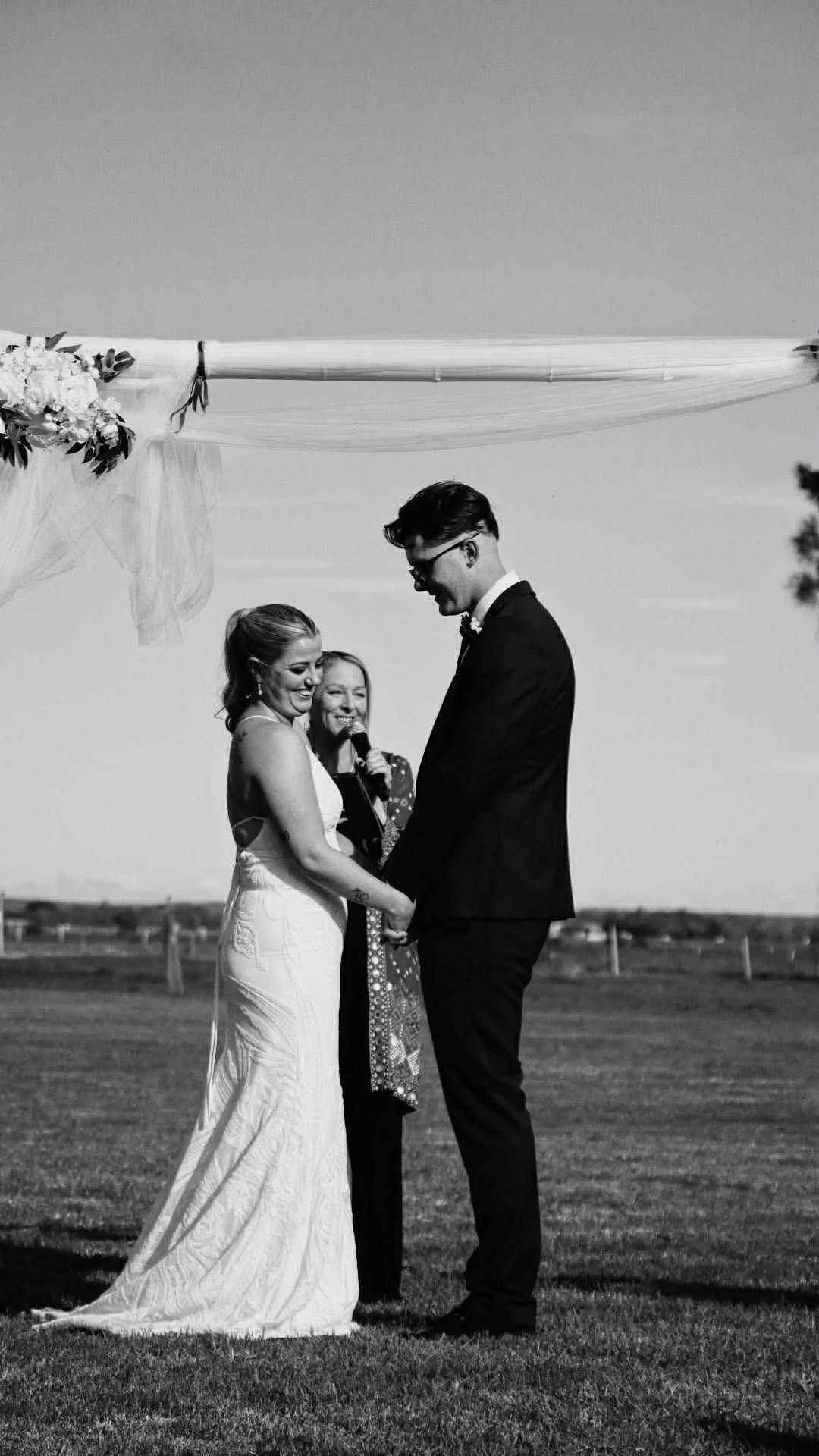 Black and white photo of a bride and groom holding hands, standing outdoors on grass, smiling at each other during a wedding ceremony, with a woman officiant in the background.