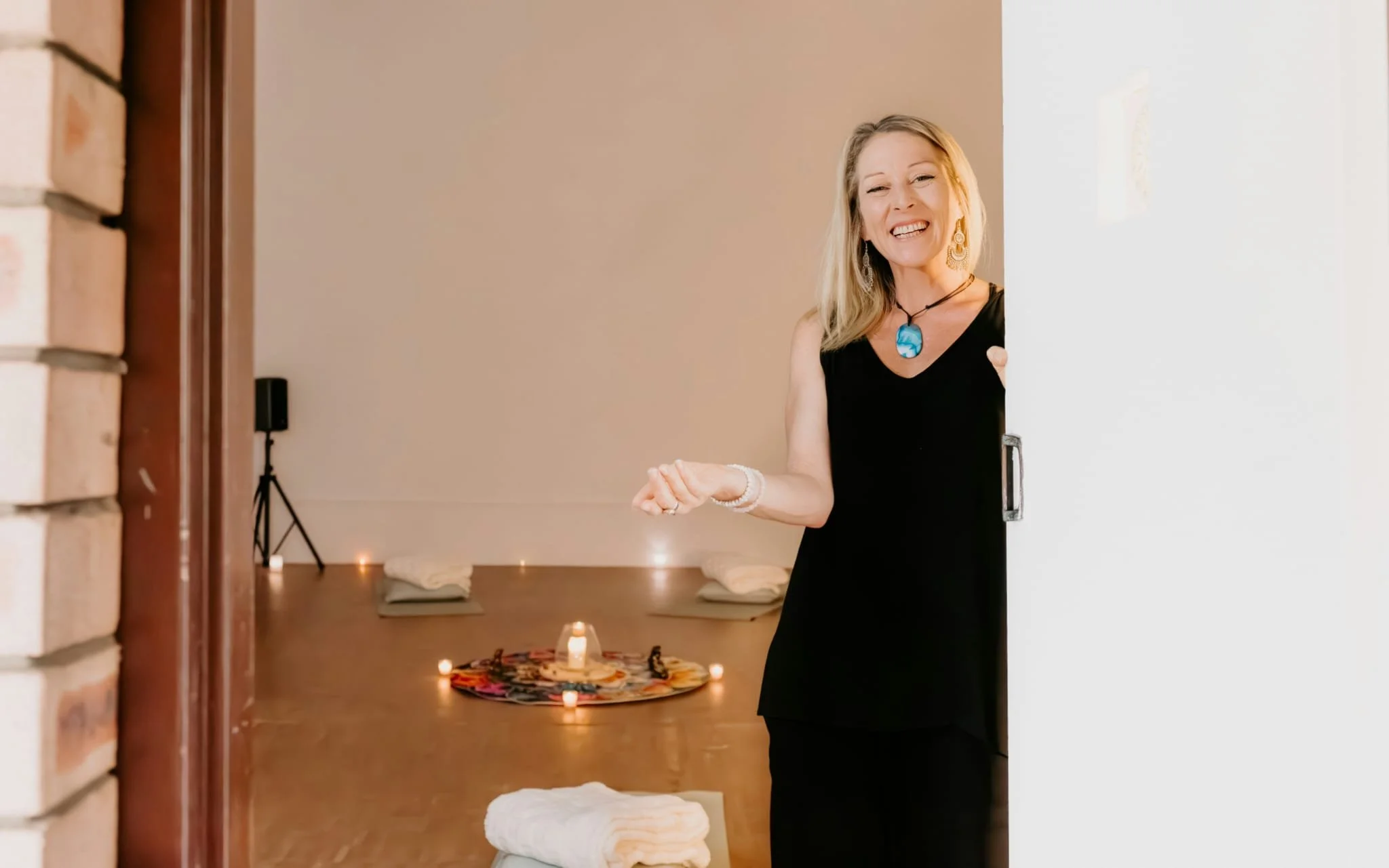 A woman in a black dress smiles as she observes a table with candles and a colorful cloth in a room set up for relaxation or meditation.