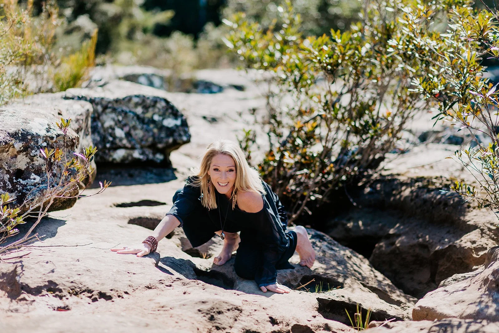 A woman with blonde hair, wearing a black outfit, is climbing on rocks outdoors among bushes and trees. She is smiling and looking at the camera.