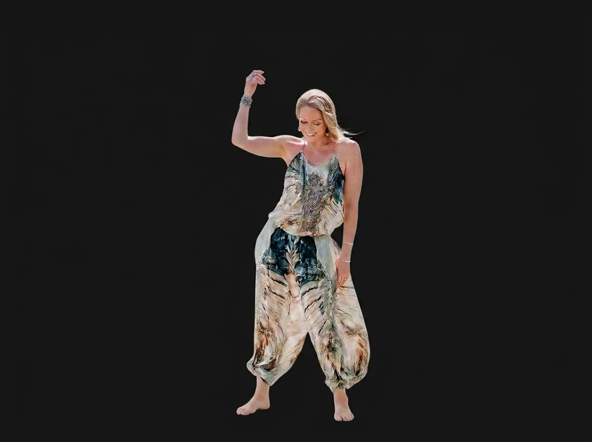 Woman dancing on a black background, wearing a colorful, flowing jumpsuit and barefoot, smiling and with one arm raised.