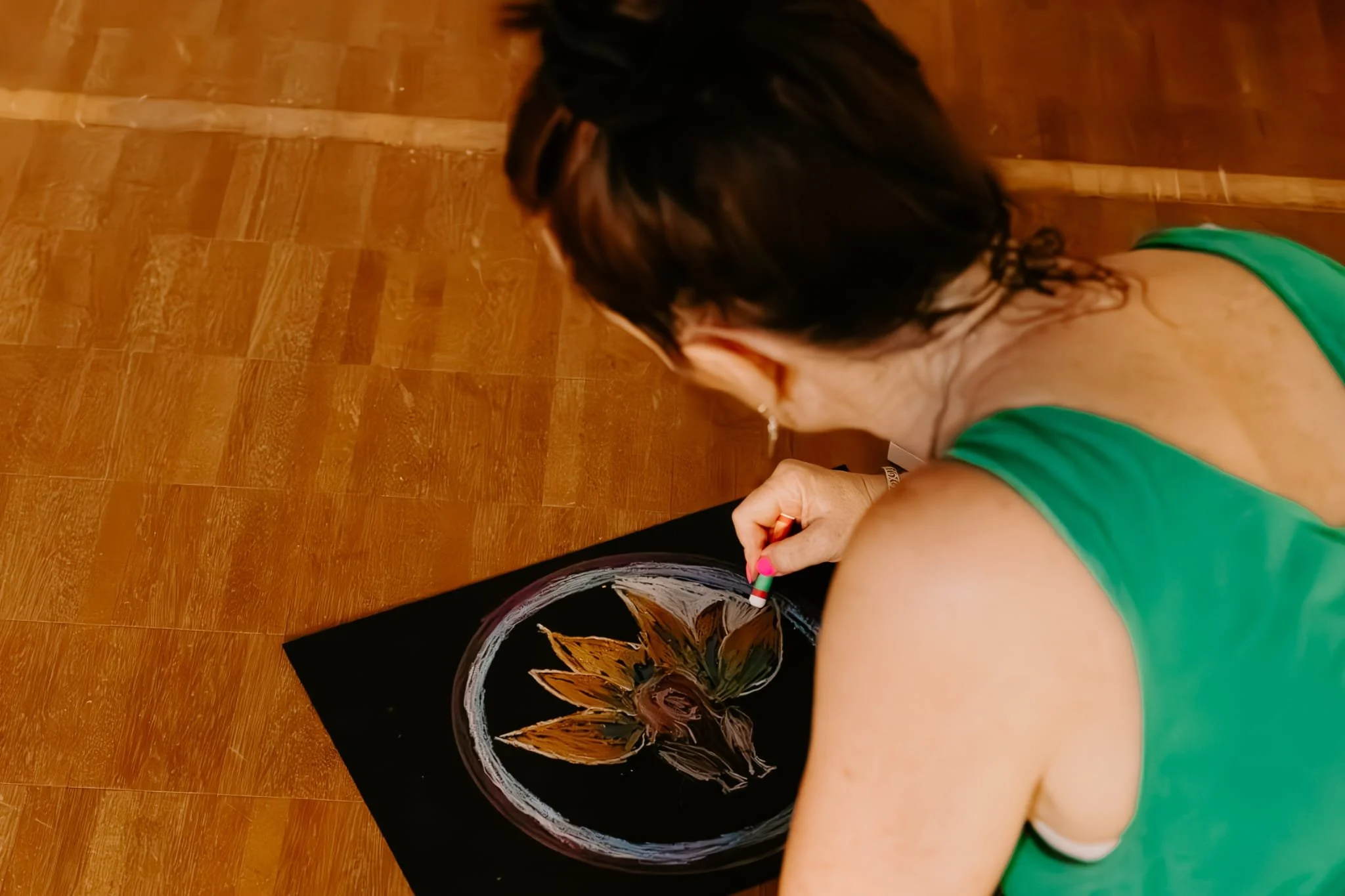 A woman with black, wavy hair and earrings is decorating a black pumpkin with white and orange designs, using a paintbrush, on a wooden floor.