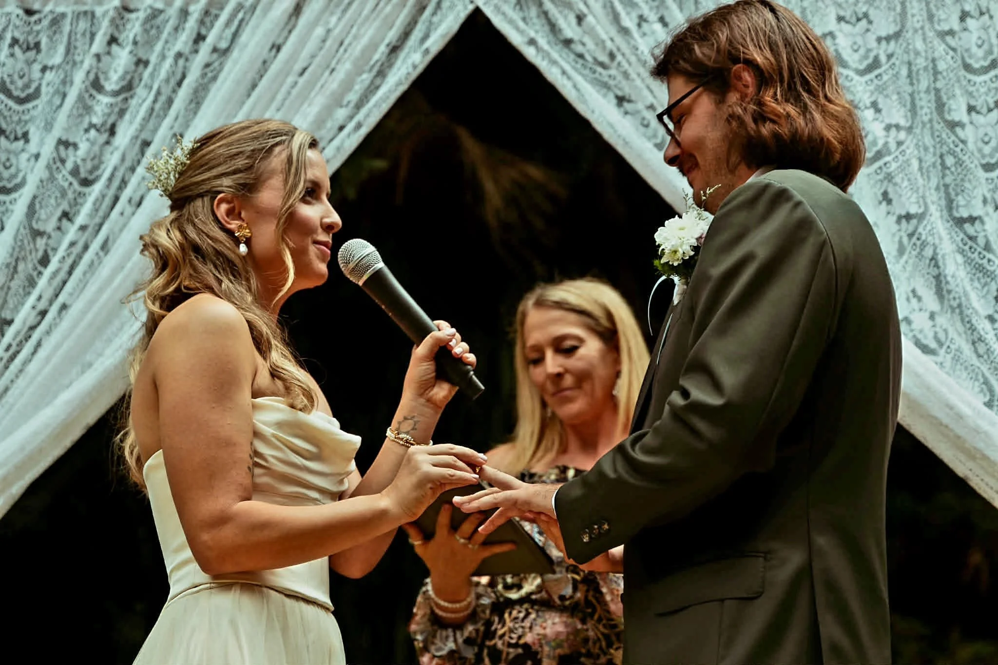 A bride and groom exchange wedding rings during their ceremony in front of an officiant, with a lace curtain backdrop.