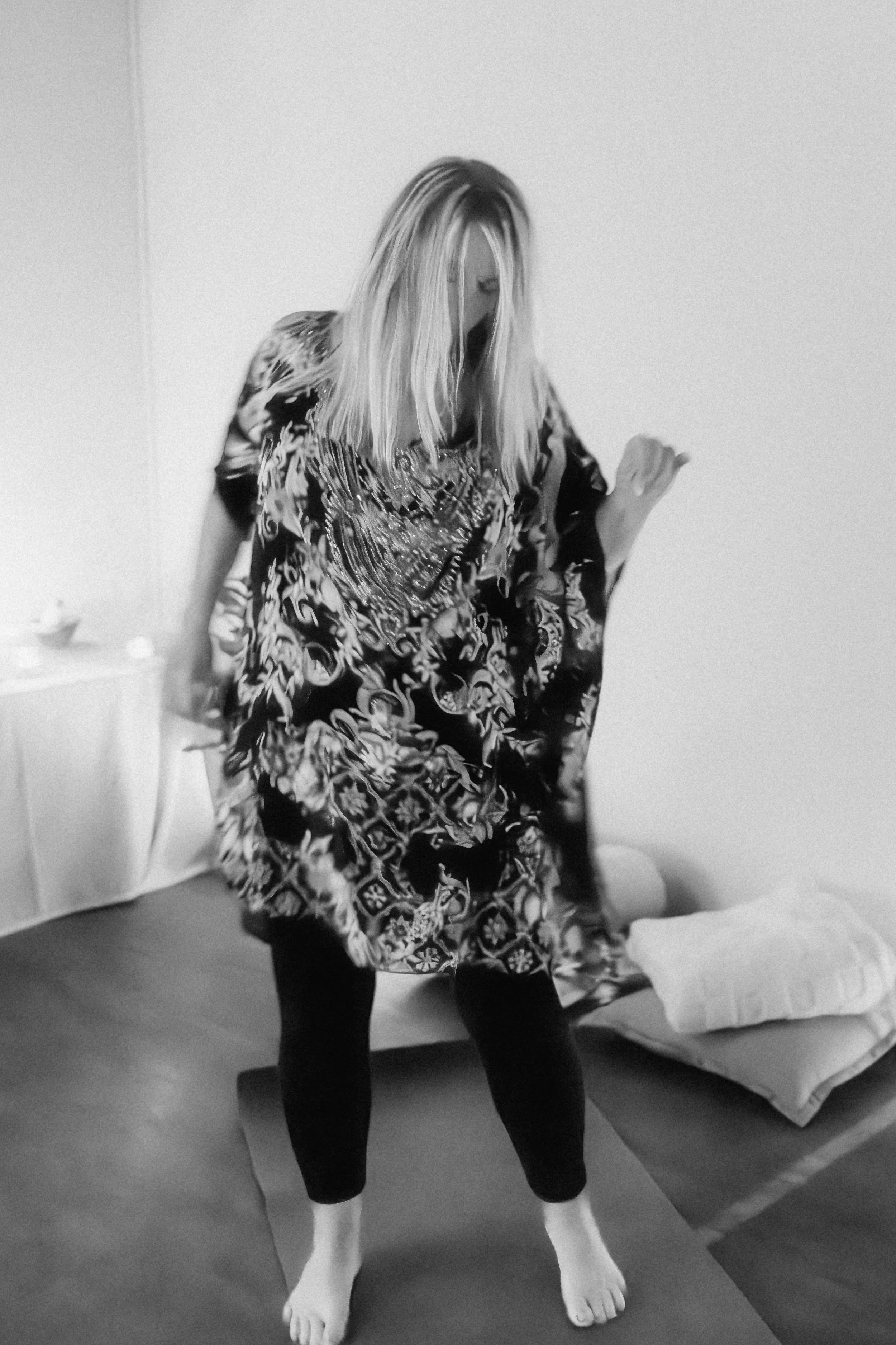 A woman with long blonde hair kneeling on a yoga mat in a room, wearing a patterned robe, with a bed and a small table visible in the background.