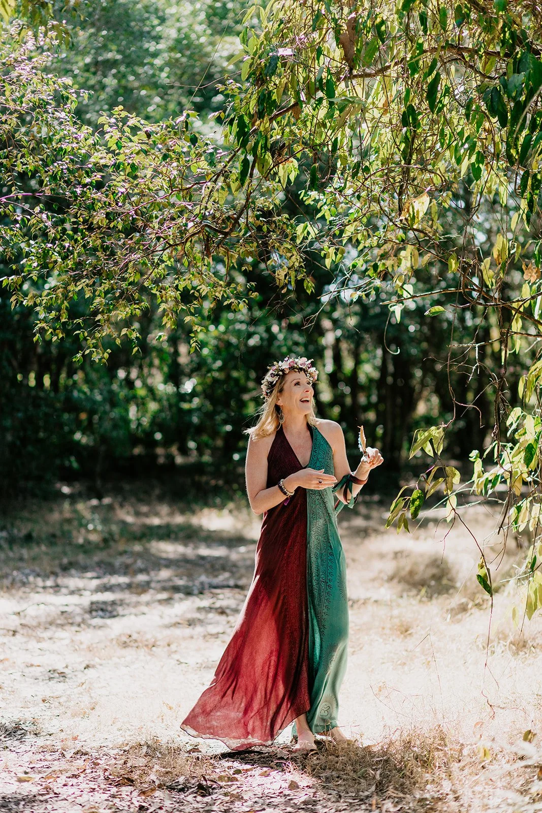 A woman wearing a colorful dress and floral crown walking outdoors in a wooded area, holding a butterfly.