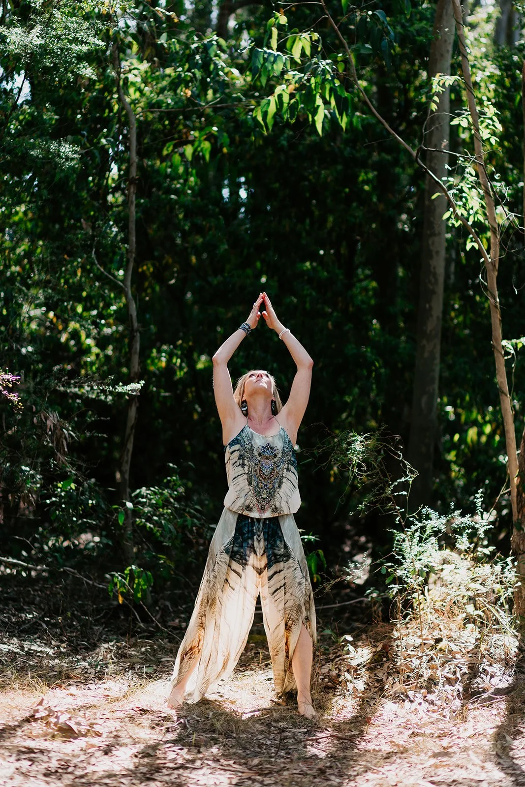 A woman standing barefoot in a forest, with her arms raised above her head, looking upward. She is wearing a sleeveless dress with animal print patterns and jewelry, surrounded by green trees and sunlight filtering through the leaves.