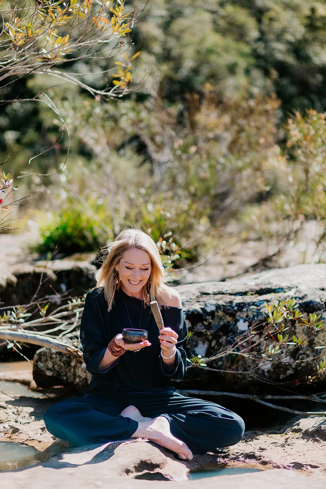 A smiling woman sitting cross-legged on the rocks by a stream in a natural outdoor setting, holding a singing bowl and a wooden mallet, surrounded by greenery.