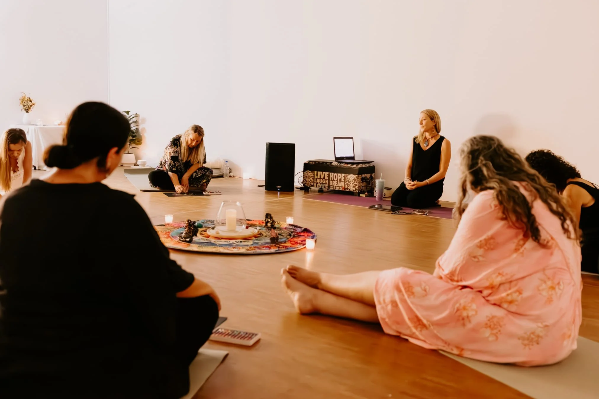 People sitting cross-legged on the floor in a circle during a meditation or yoga session in a bright, minimalistic room with white walls and wooden flooring.