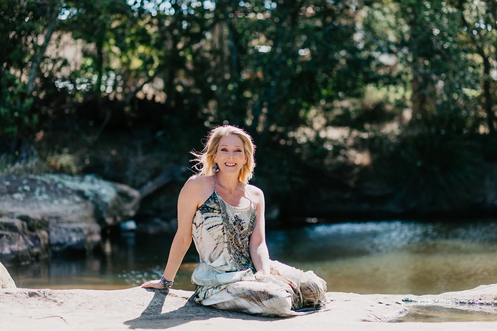 A woman with blonde hair sitting on a rock by a river, smiling, wearing a patterned dress with jewelry, surrounded by lush green trees.
