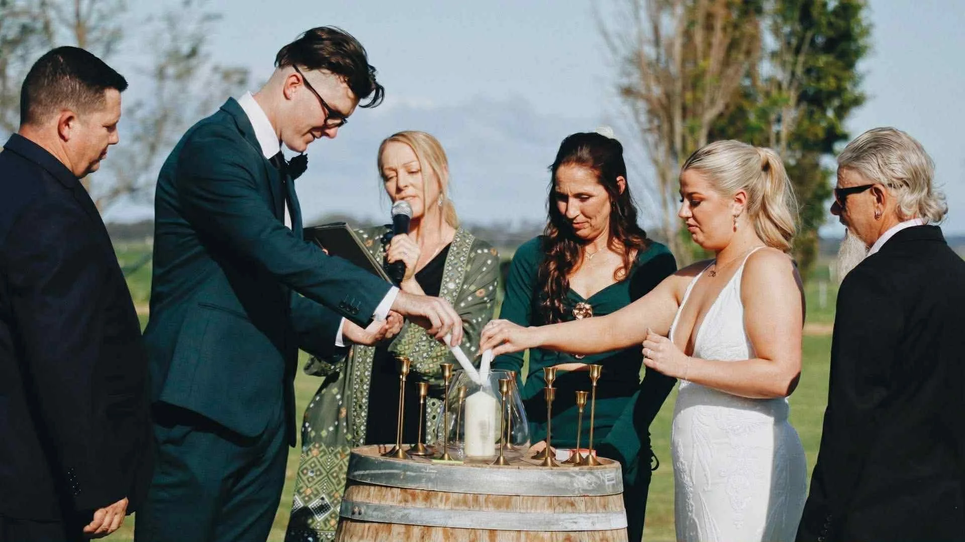 A wedding ceremony outdoors, with a bride and groom lighting a unity candle while officiants and guests watch.