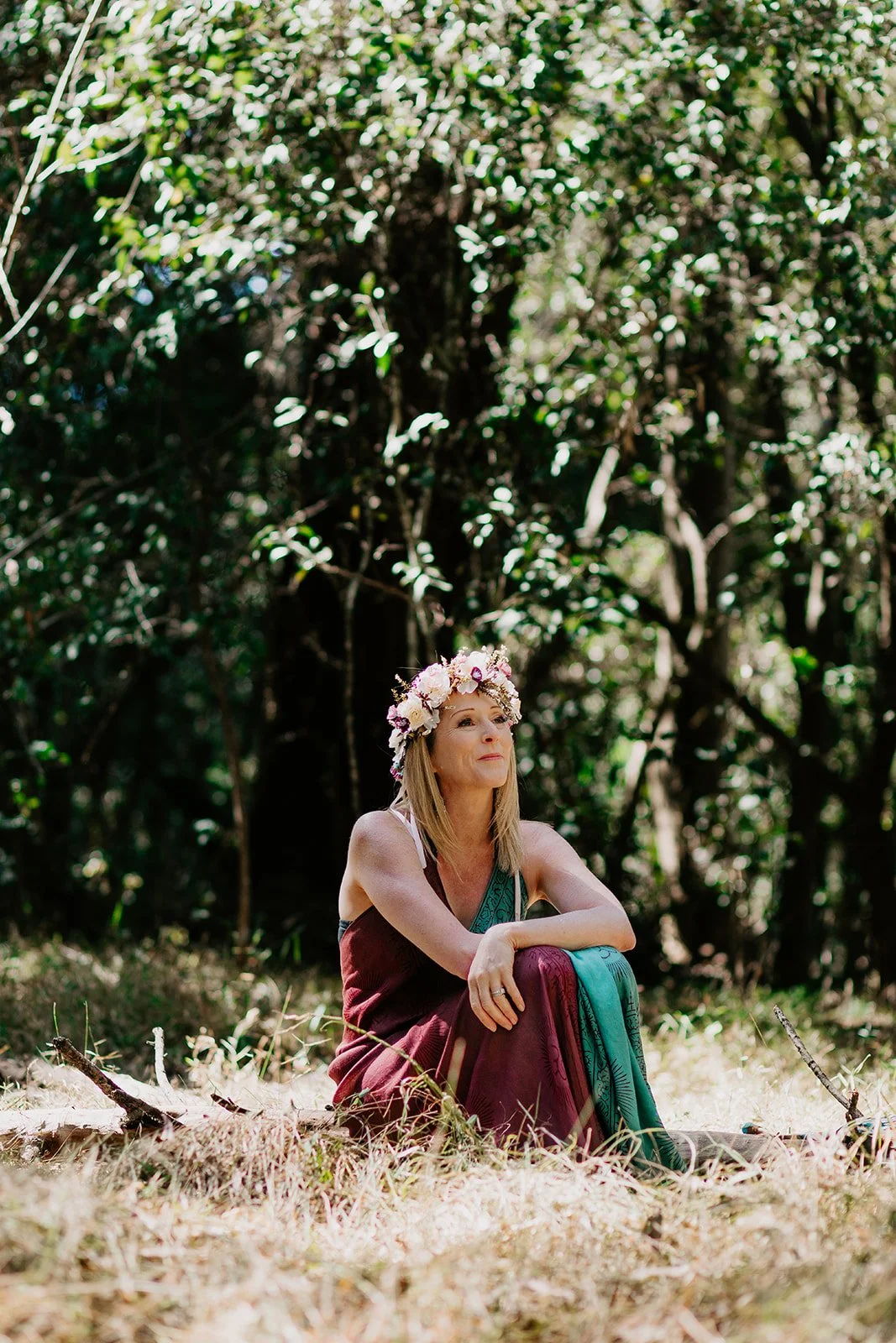 A woman wearing a flower crown sitting outdoors in a forested area with sunlight filtering through trees.