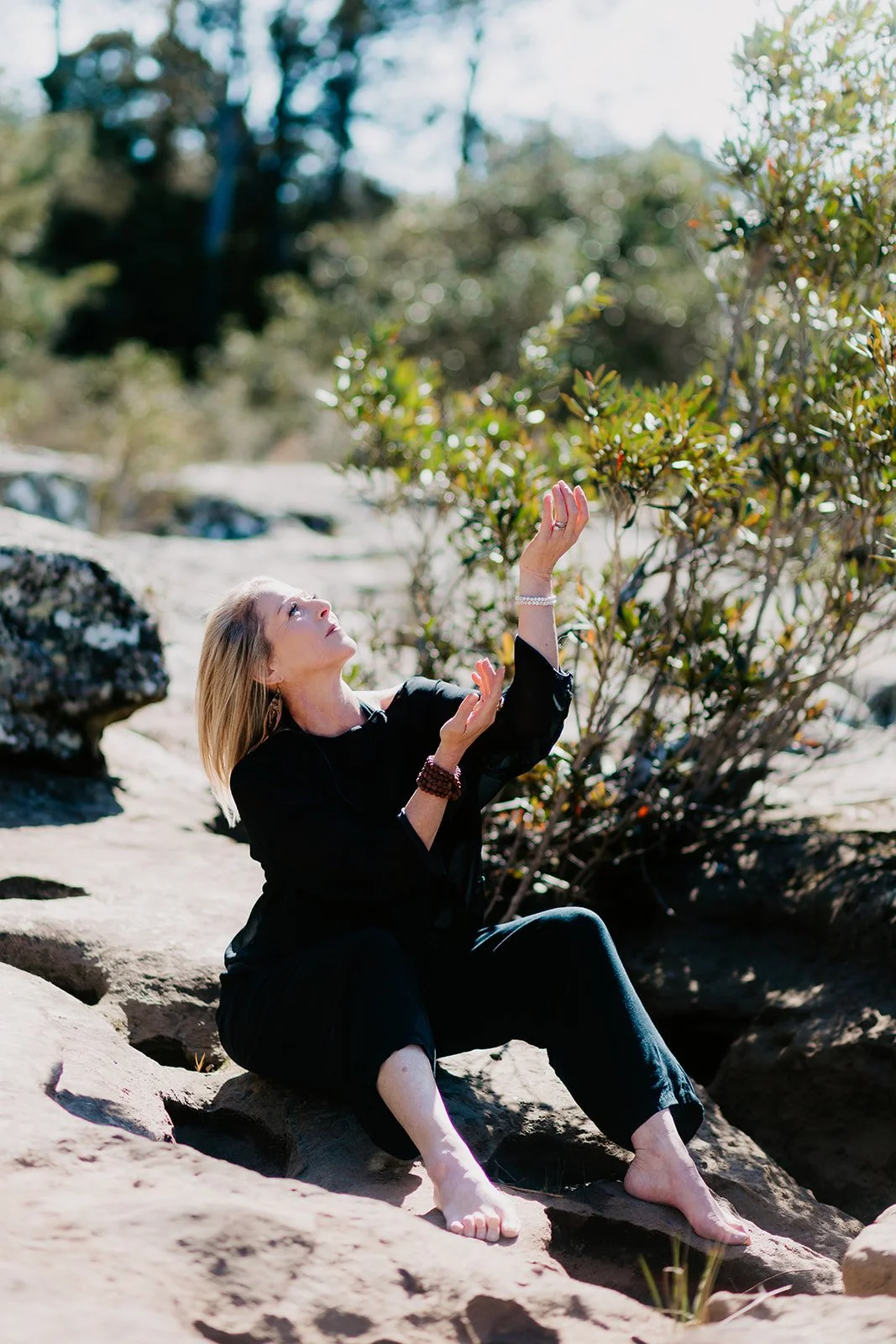 A woman sitting on rocks outdoors, reaching up to a tree branch, with greenery and rocks in the background on a sunny day.