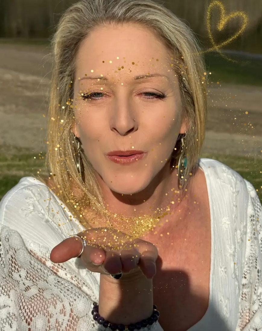 A woman blowing golden glitter from her hand outdoors, with a sparkly heart above her head.