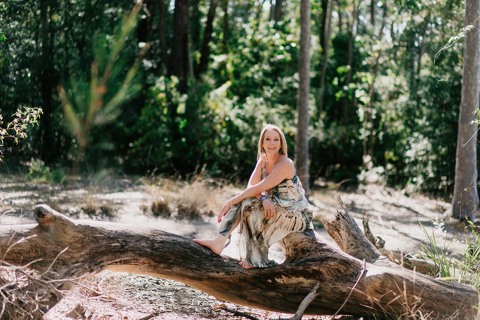 A woman with long blonde hair sitting on a fallen tree trunk in a forest, wearing a flowy dress with animal print, smiling at the camera with trees and sunlight in the background.