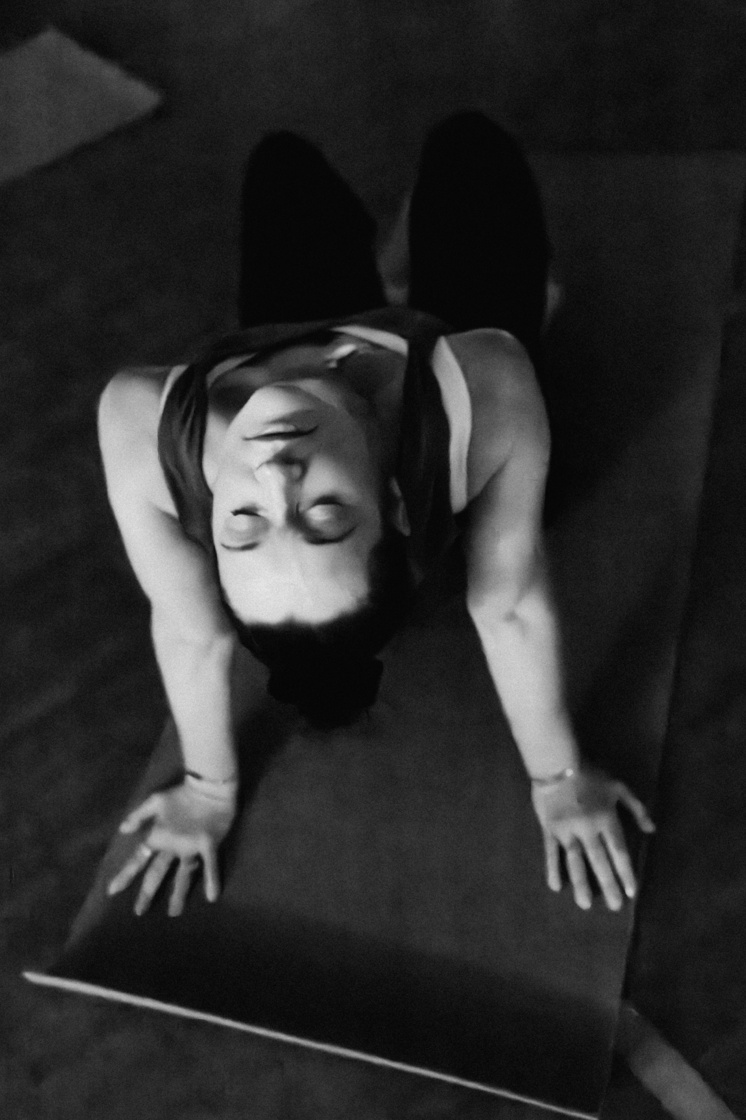 A person with long hair and glasses, wearing a sleeveless top, performing a yoga pose on a dark surface with their hands and forehead touching the ground, in black and white.