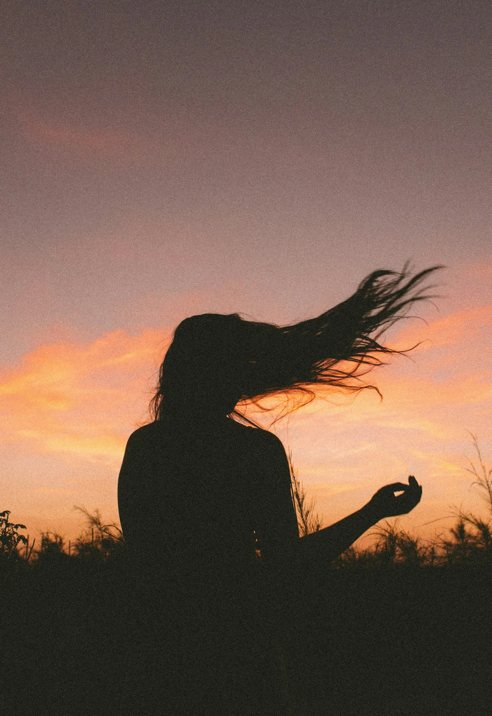 Silhouette of a person with long hair blowing in the wind, sitting or kneeling outdoors during sunset with a colorful sky and clouds in the background.