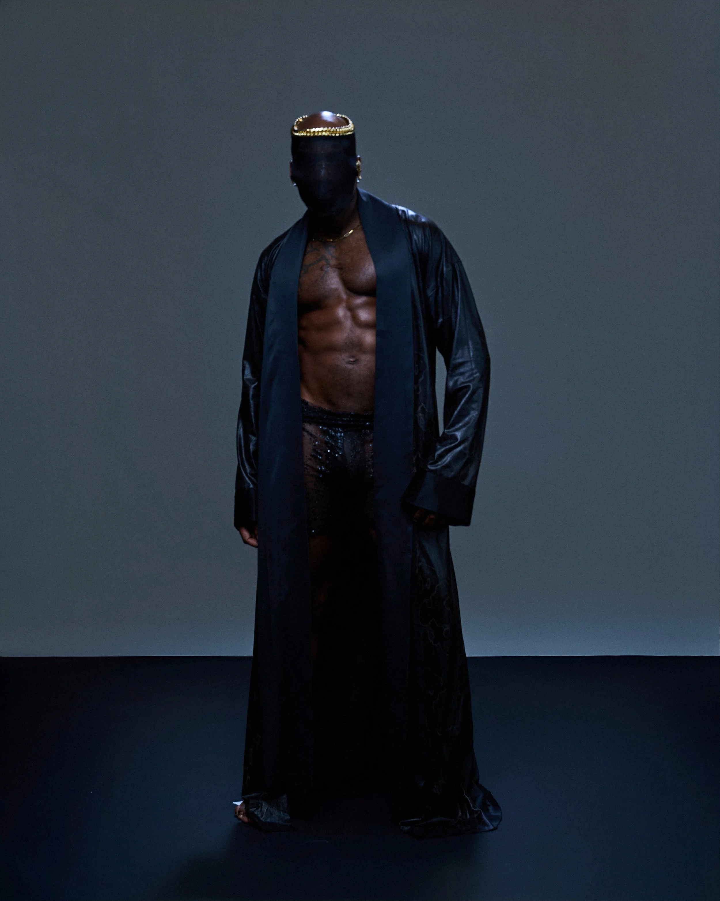 Geoffrey Smith - A person with a dark face mask, no shirt, wearing a black open long coat, black sequined pants, and black shoes, standing against a gradient dark background.