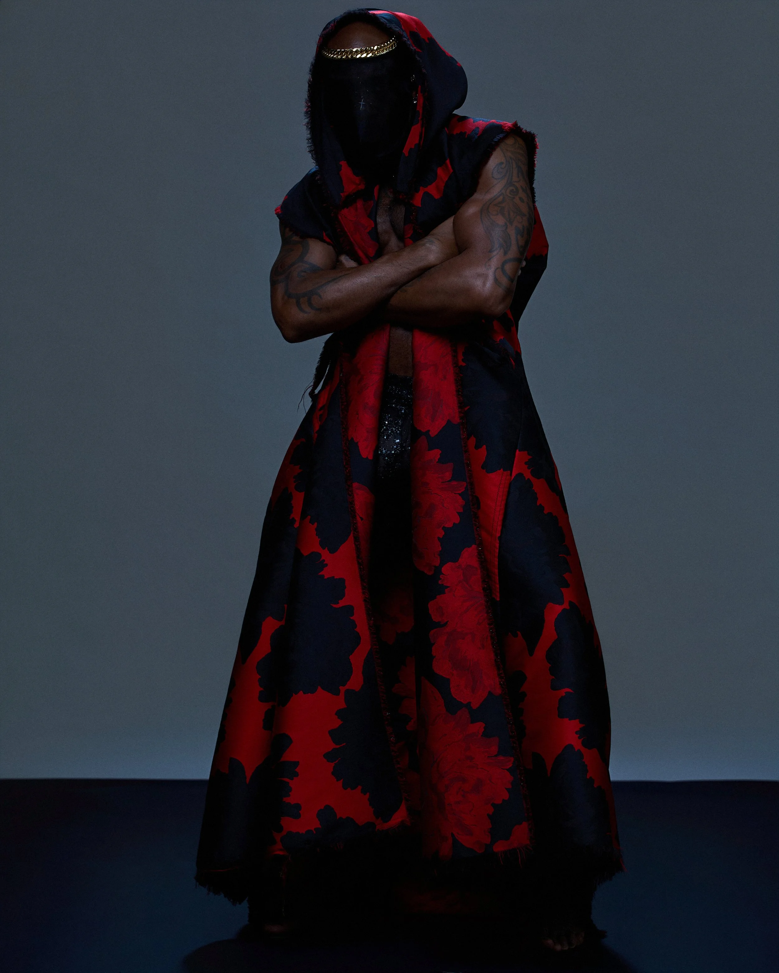 A person with dark skin, black hair, and tattoos on their arms, wearing a red and black patterned robe with a hood, a gold chain headband, and black face covering, standing with arms crossed against a plain dark background.