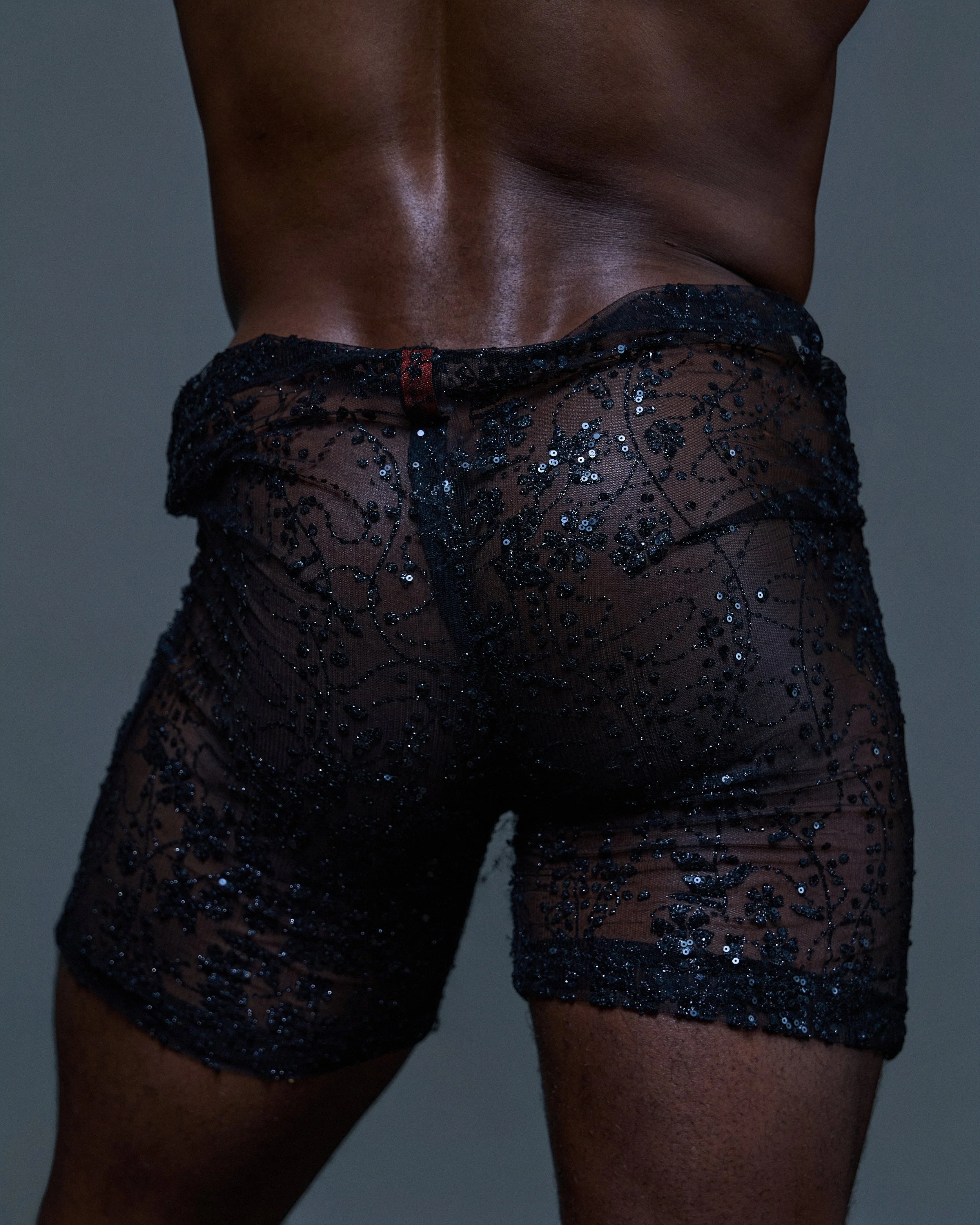 Close-up of a person's lower back and upper thighs, wearing black sheer underwear with sparkly embellishments.