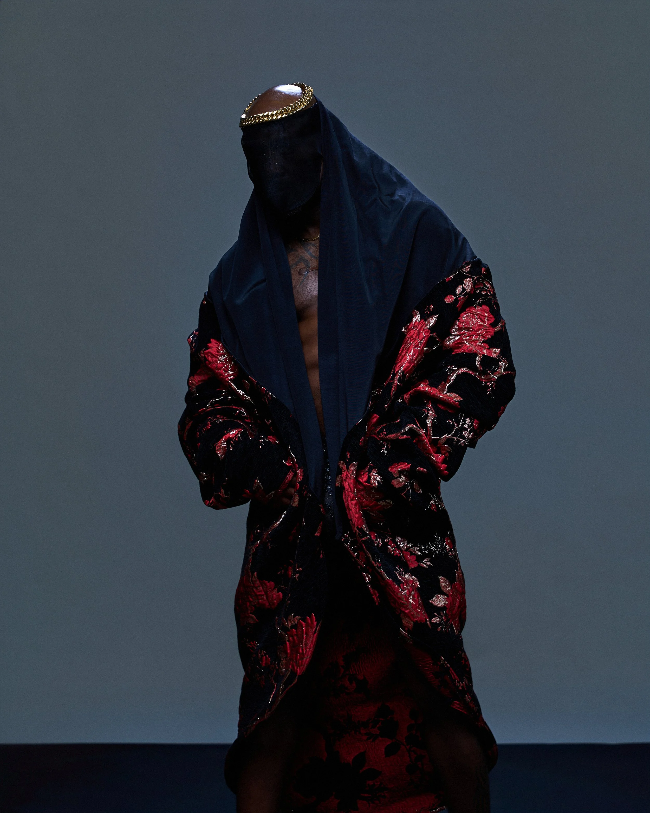 A person wearing a dark hooded cloak with a gold chain around their forehead, a black face covering, and a black and red embroidered jacket, standing in front of a gray background.