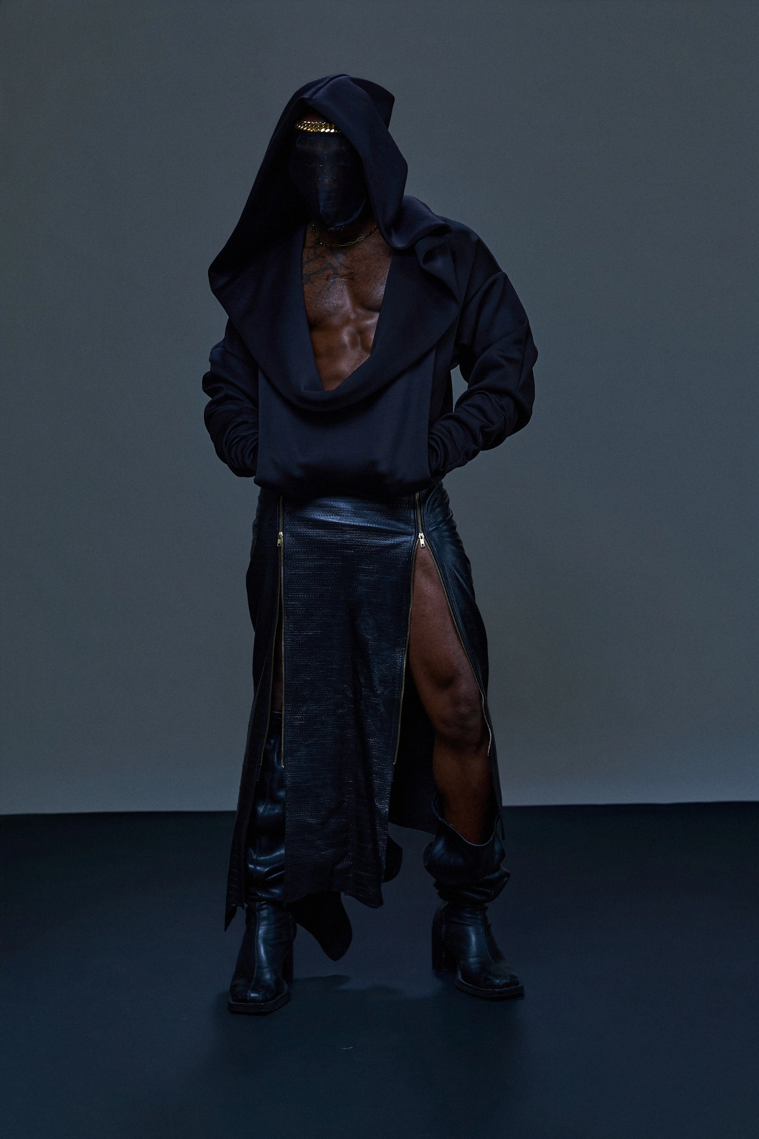 Person wearing black leather boots, a long black leather skirt with a high slit, and a black hoodie with the hood up. The person's face is covered with a black mask and they are standing against a dark background with hands in the pockets of the hoodie.