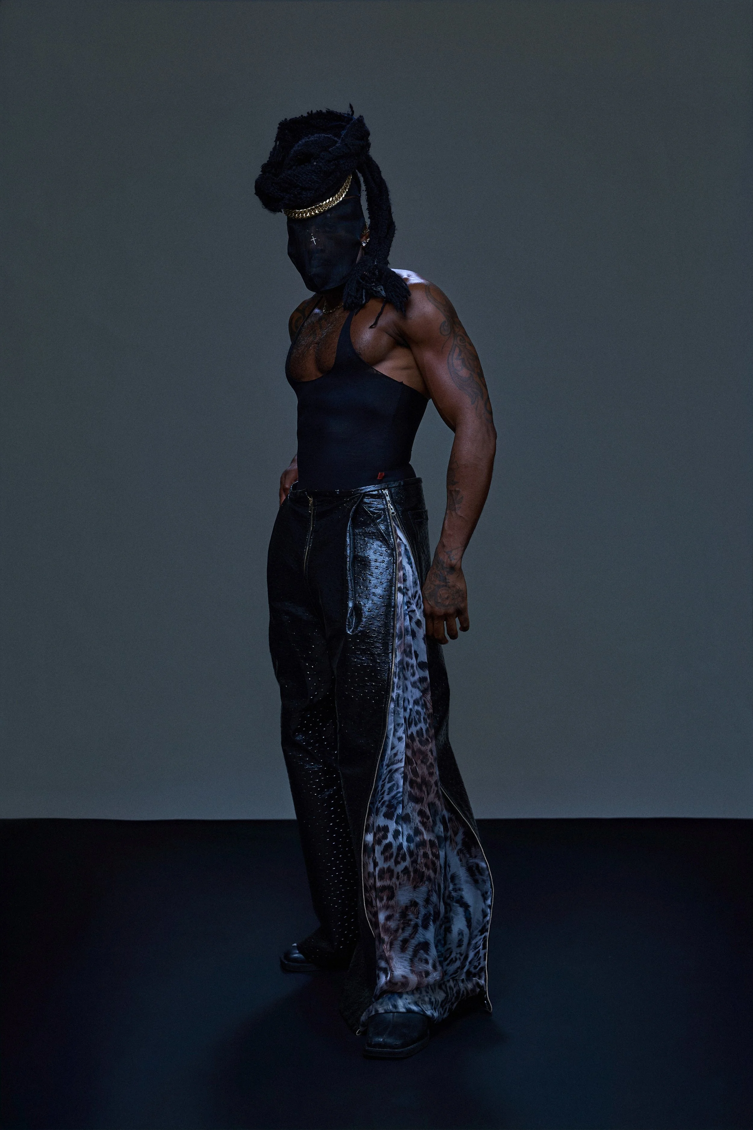 A person with long dreadlocks wearing a black mask, black sleeveless top, black shiny pants, and black boots stands against a dark background.