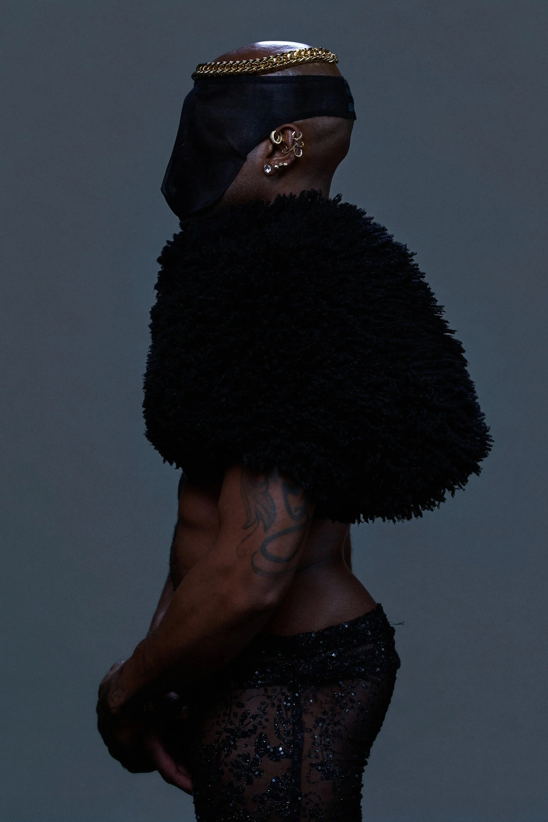 A person with dark skin, visible tattoos on their arm, wearing a black face covering, gold chain headband, multiple earrings, and dark clothing with glittering details, is standing against a plain gray background.