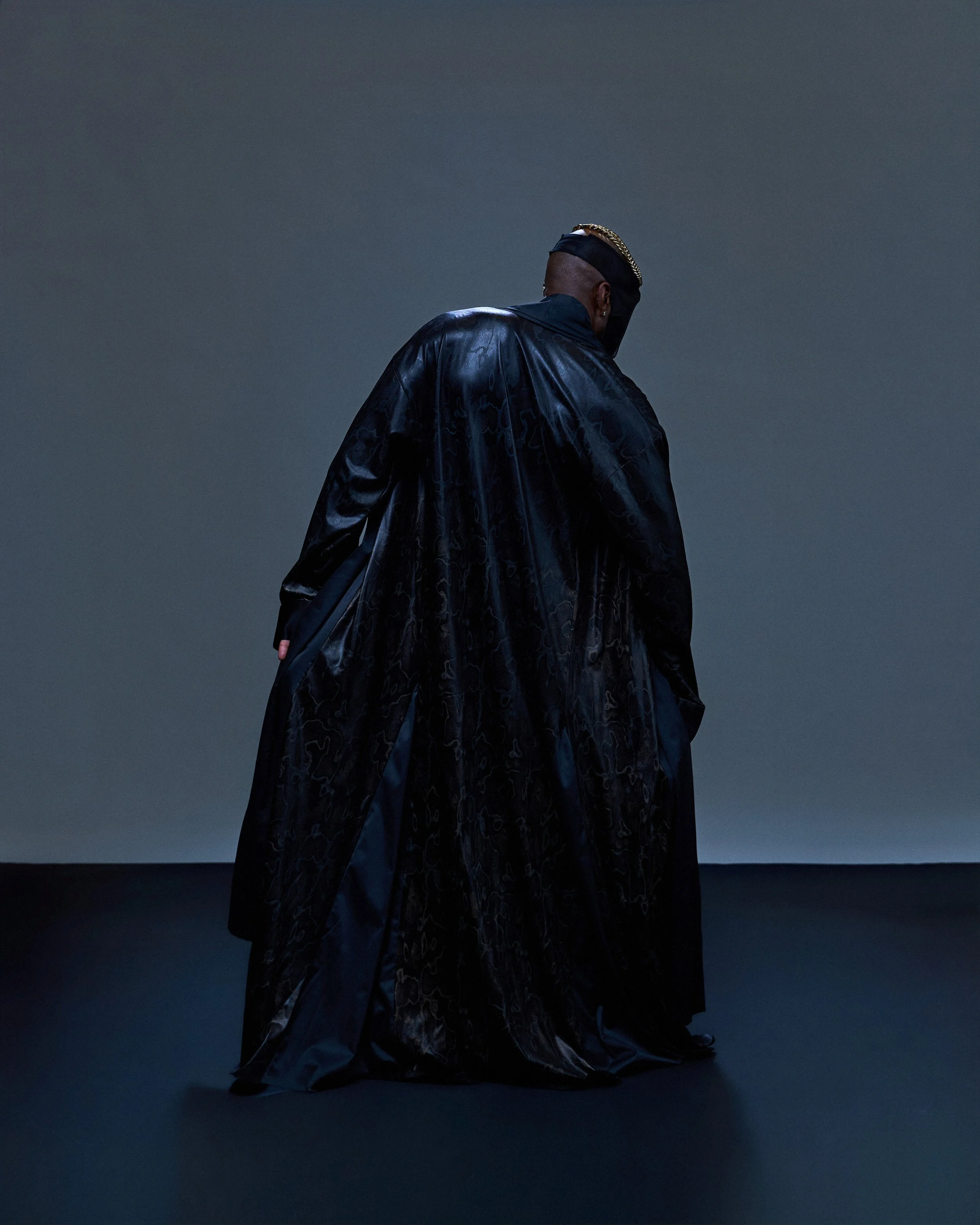 Geoffrey Smith - Person in black clothing with a shiny, textured appearance stands against a dark, gradient background, facing away from the camera.