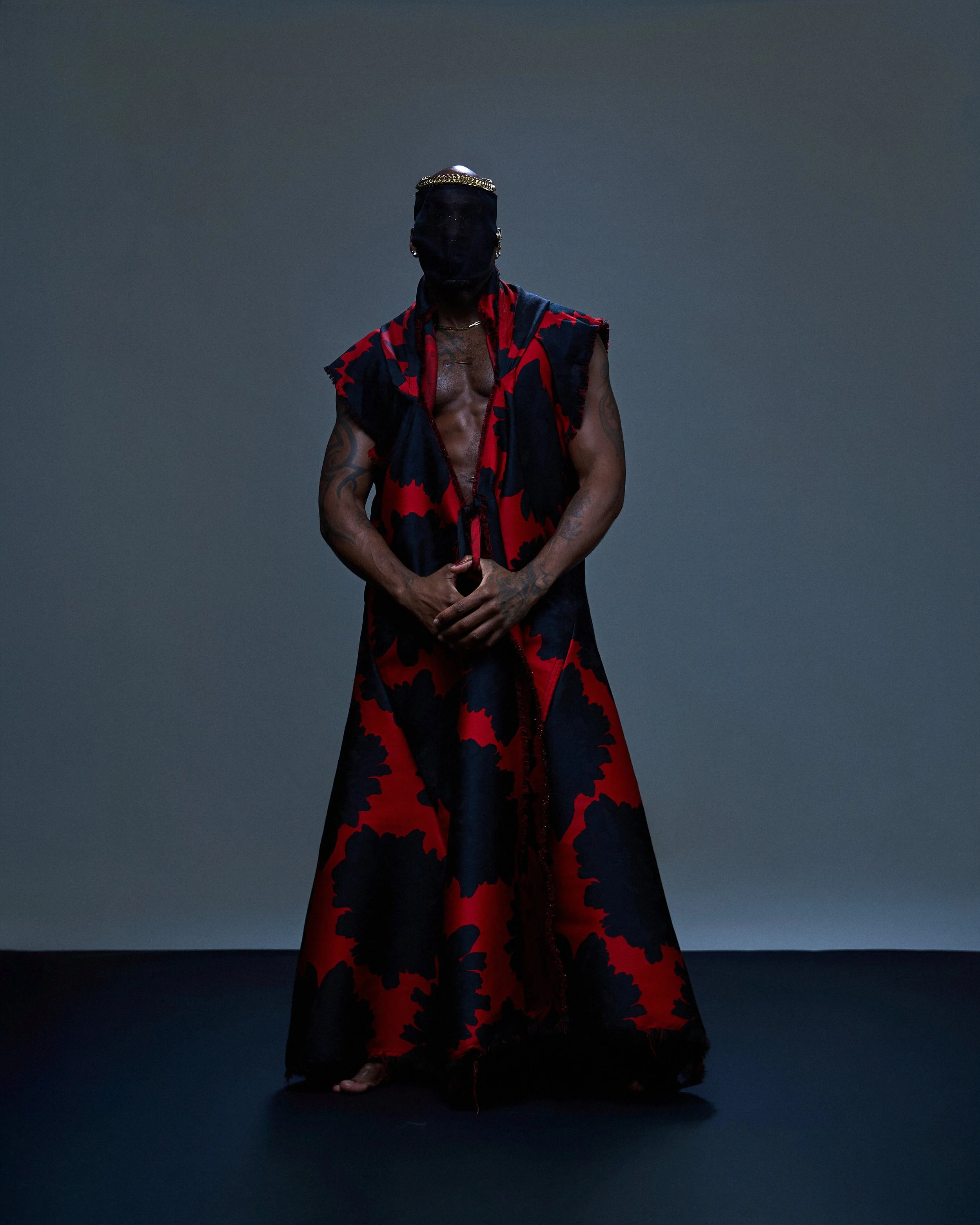 Person standing in front of a dark gray background, wearing a faceless black mask, torn red and black clothing, with tattoos on arms and visible chest, holding their hands together.