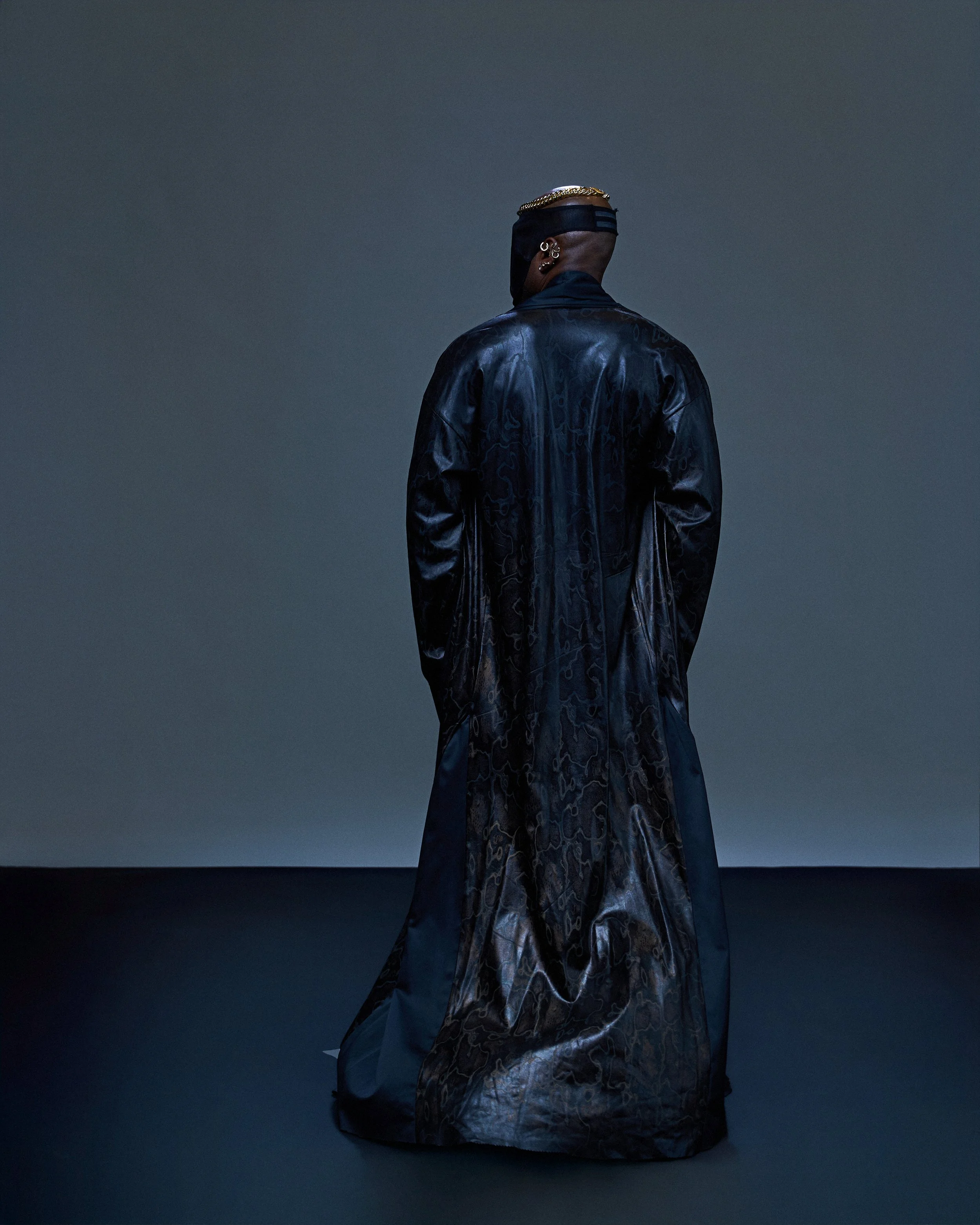Geoffrey Smith - Rear view of a person wearing a black leather trench coat, with earrings and a head covering, set against a dark, plain background.