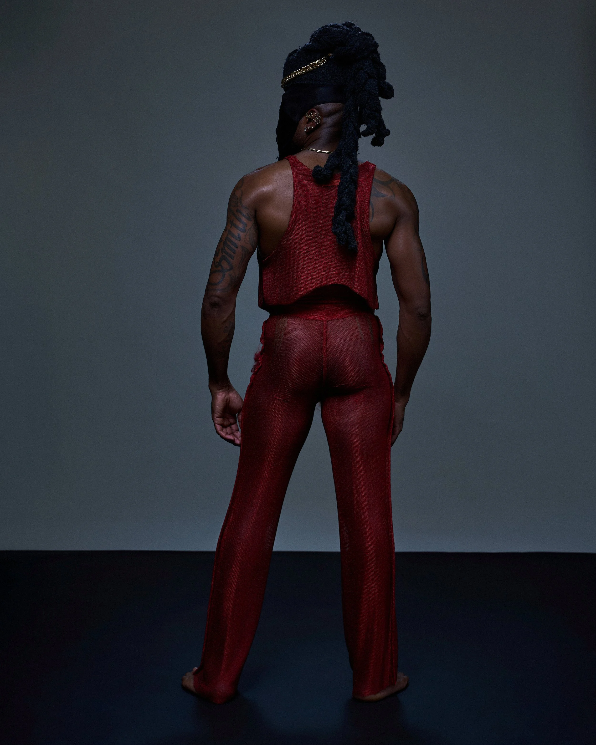 A person with dark hair styled in dreadlocks, dressed in a red sleeveless top and matching red pants, stands with back facing the camera, wearing multiple earrings and a necklace, against a dark gray background.