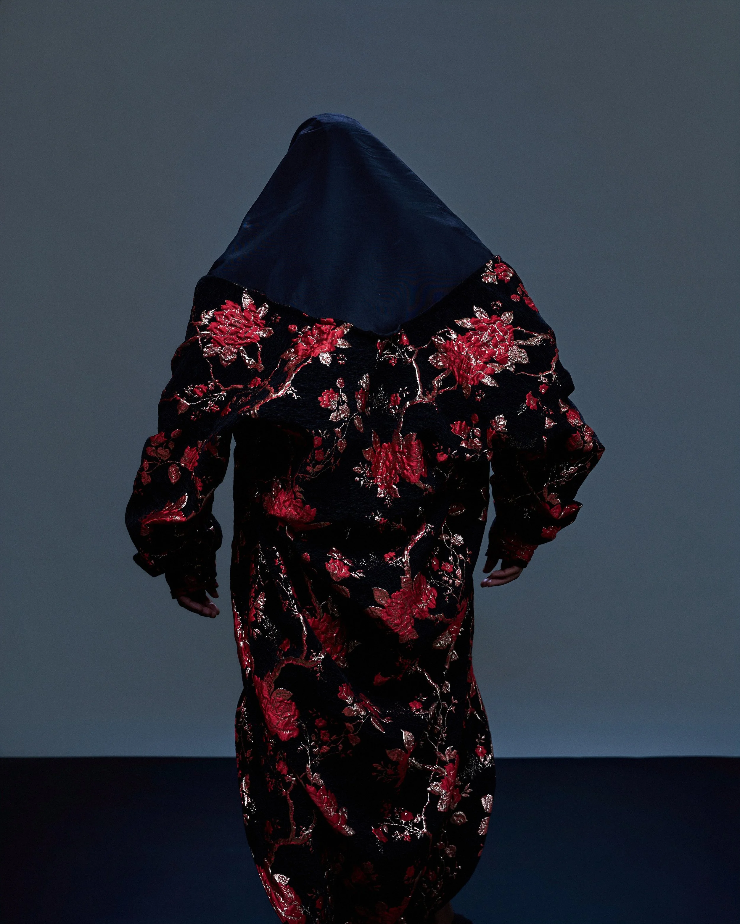 Person wearing a black dress with red and silver floral embroidery, with a dark head covering, standing against a gray background.