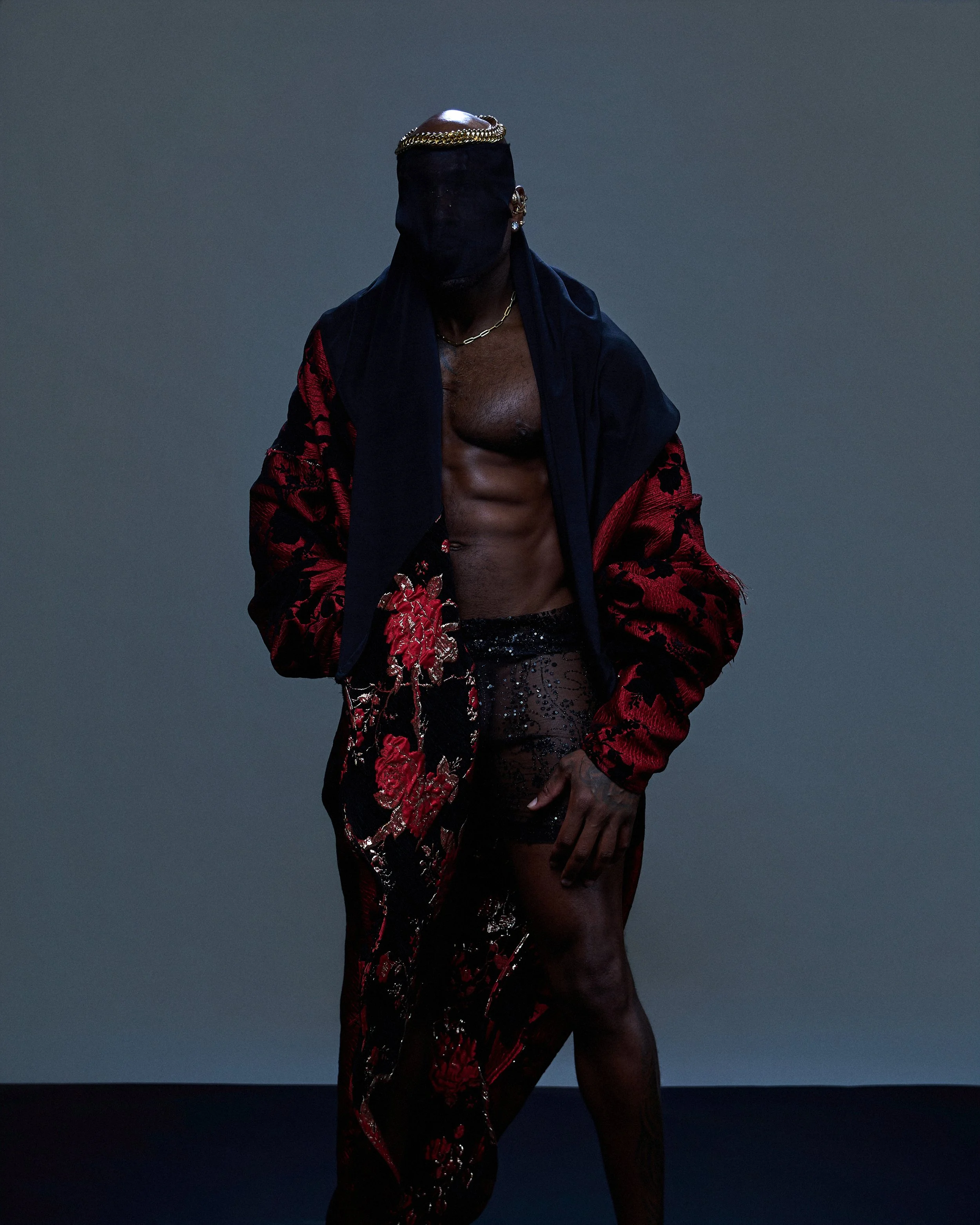 A man with a bandaged face wearing a gold chain, a veil covering his face, a black and red embroidered jacket, sheer black pants, and no shirt, standing against a dark, gradient background.