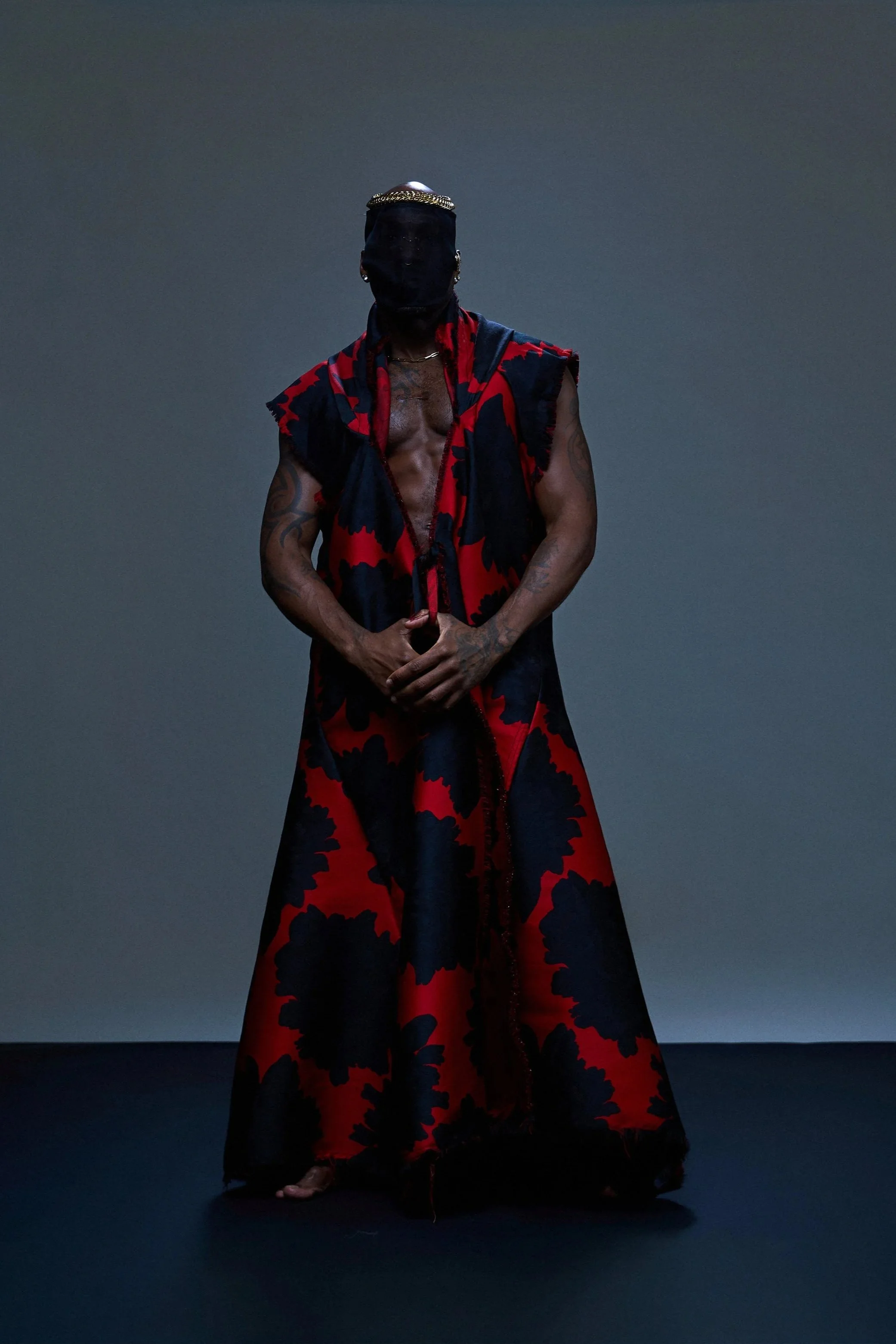 Person wearing a black face covering, jewelry, and a red and black patterned sleeveless robe, standing against a dark gray background.