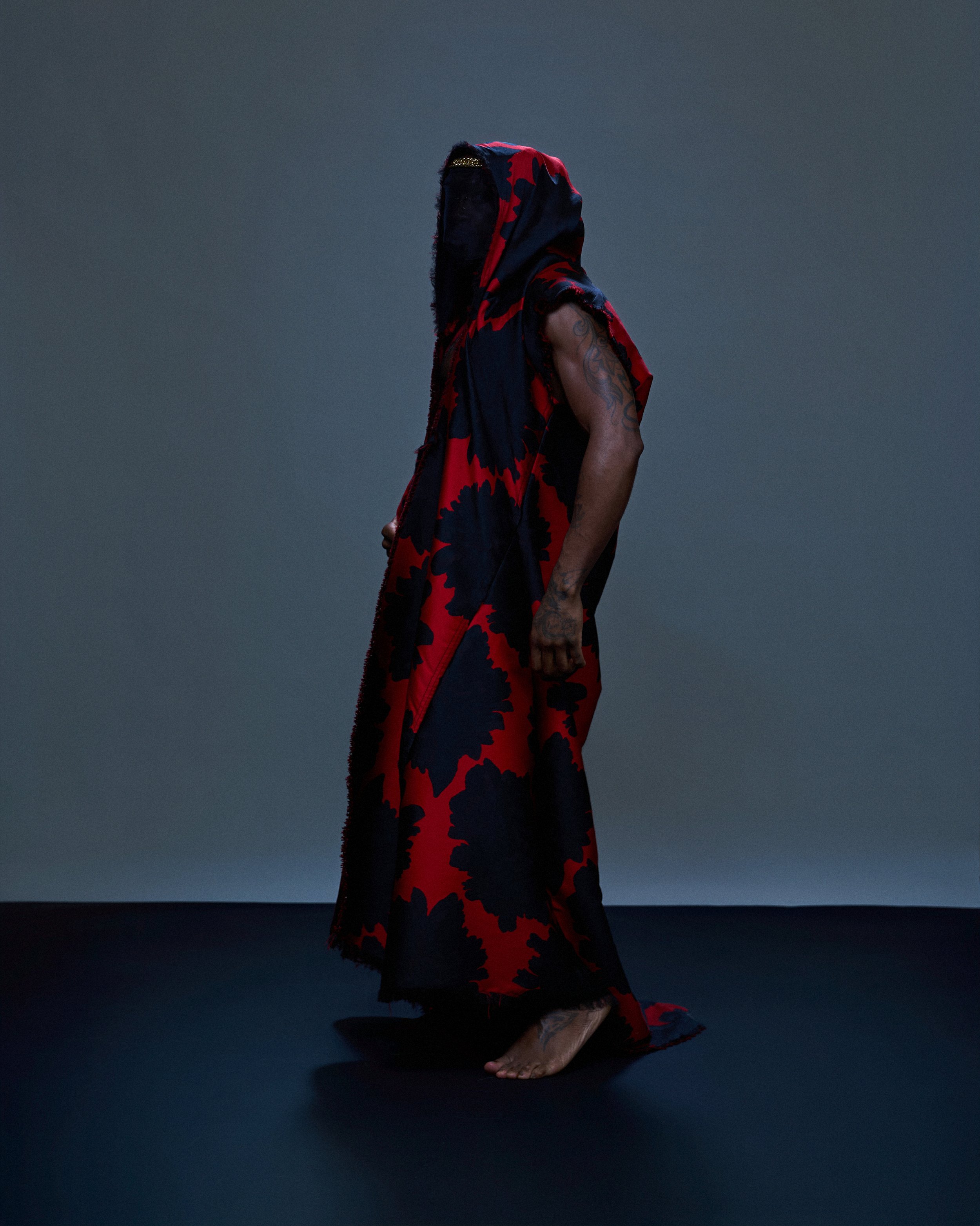 A person standing on a dark blue background wearing a black and red tie-dye full-length hooded garment, with visible tattoos on their arm, and no shoes, barefoot.