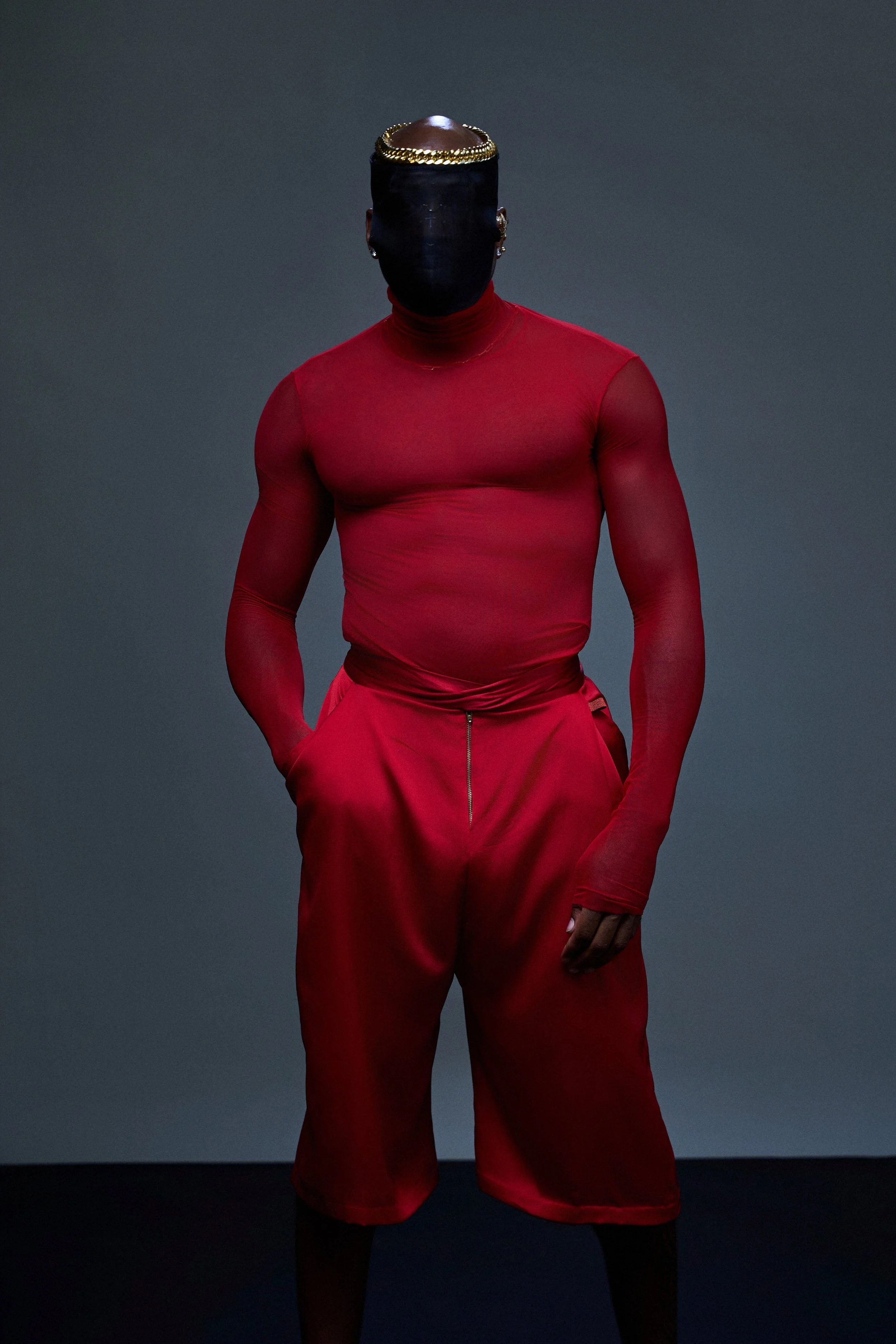 Person wearing a black face mask, a gold chain headband, a red long-sleeve shirt, and red pants against a dark background.