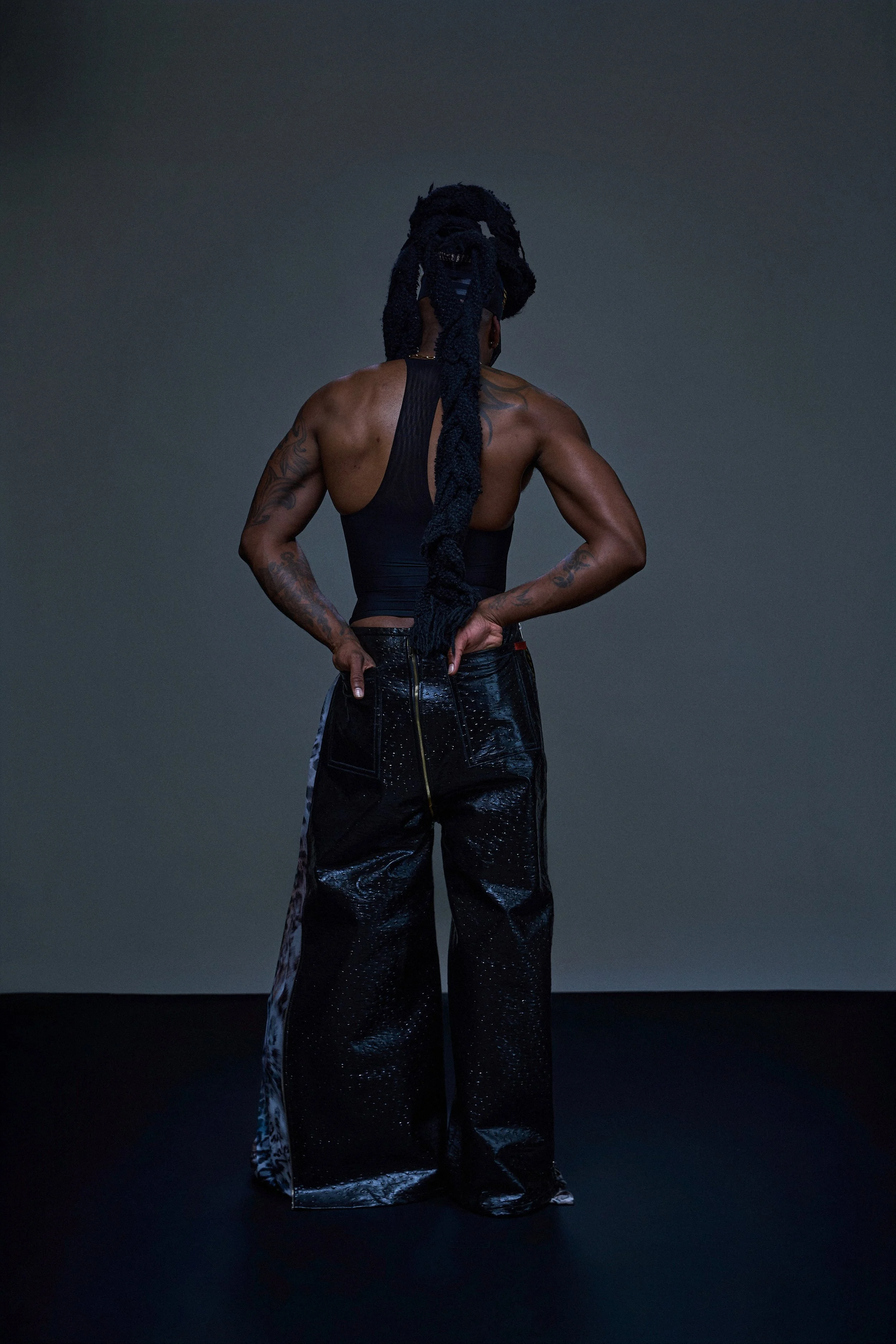 Geoffrey Smith - A person with long dreadlocks standing with their back to the camera. They have tattoos on their arms and shoulders, are wearing a sleeveless black top, and shiny black wide-leg pants, in a studio with a dark background.