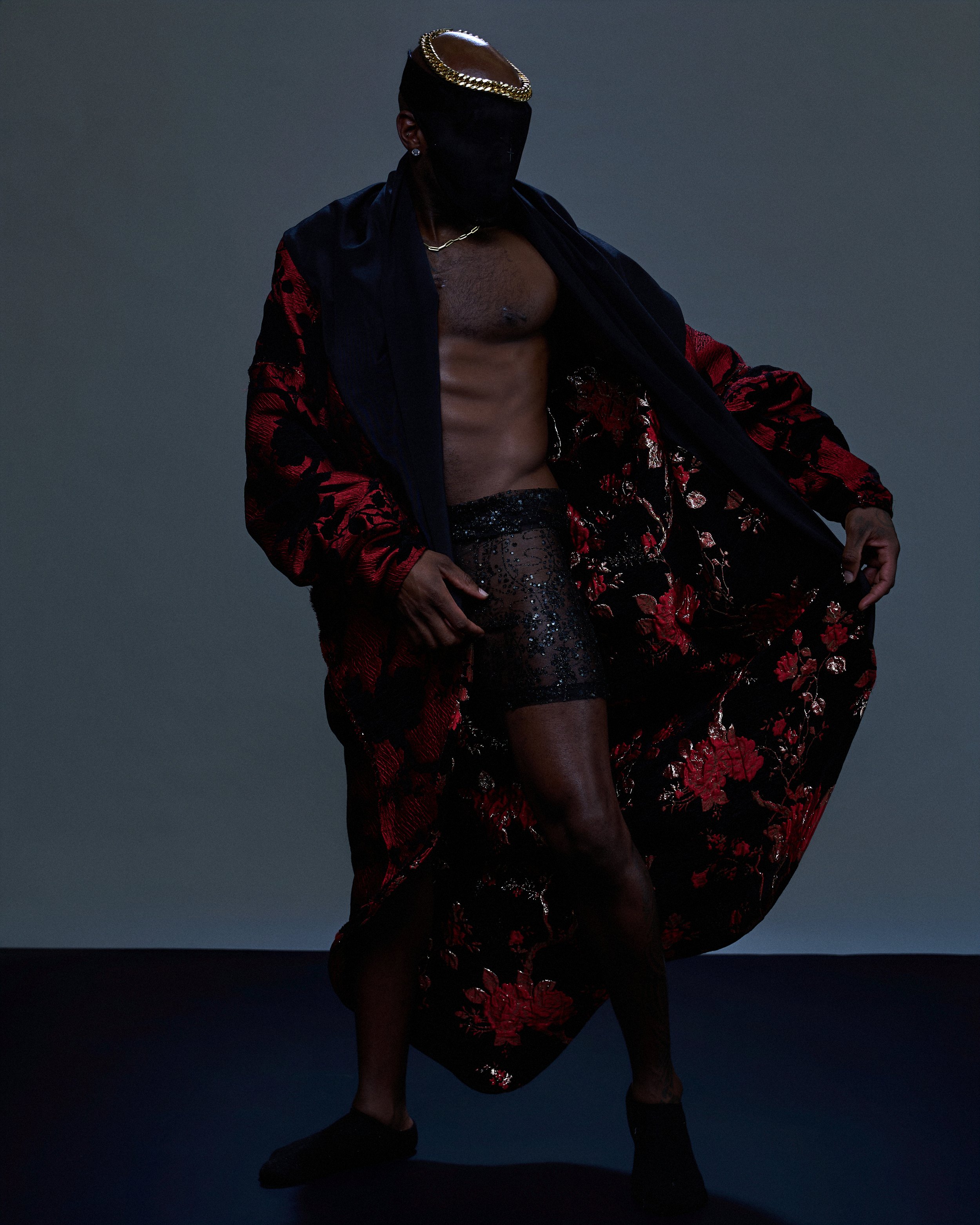 A person standing in a dark room with a gray background, wearing a black and red floral robe, black heels, and a large black face covering with gold jewelry and a leather headpiece.