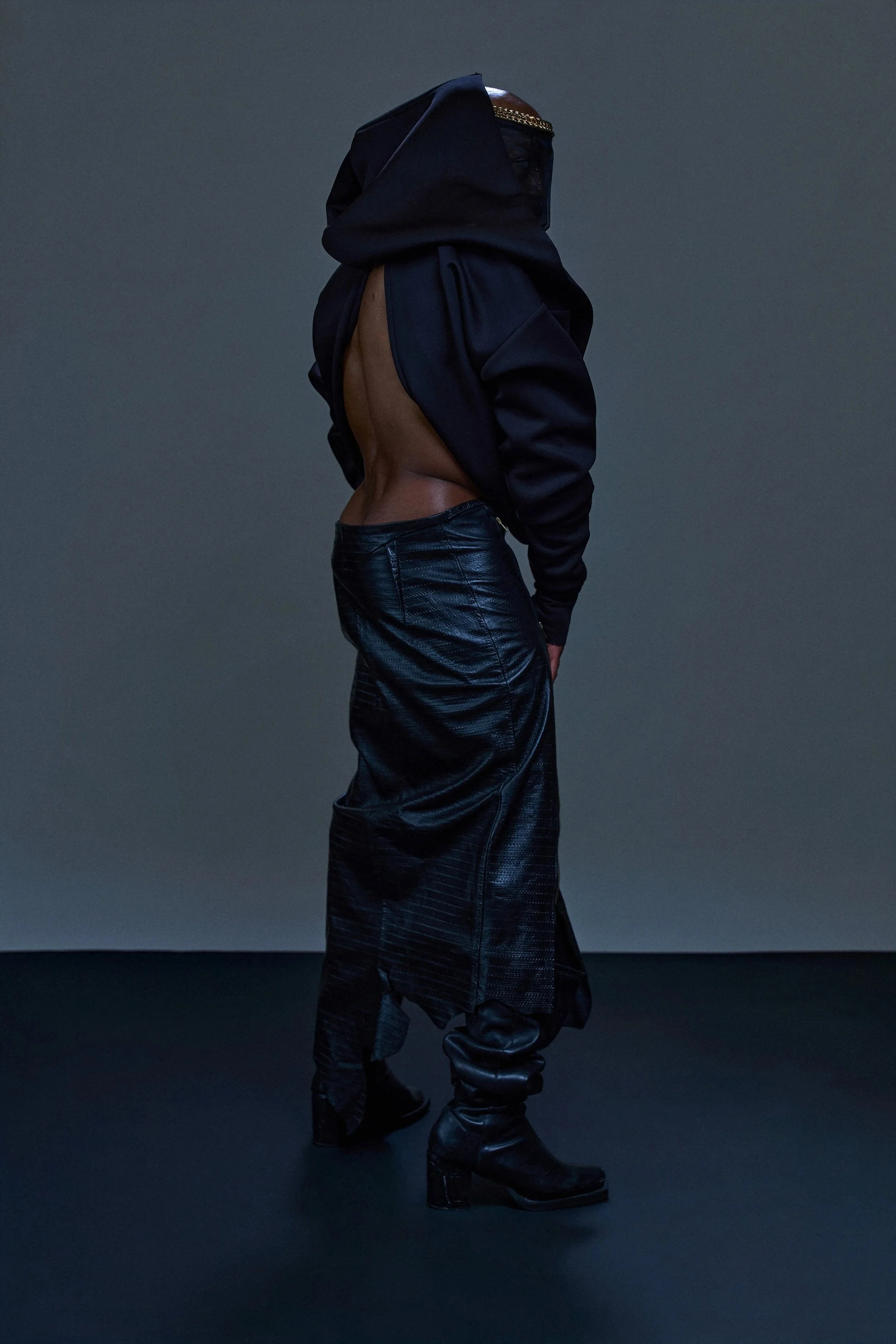 Person in black leather pants, black boots, with a large black jacket covering their face and head, standing in a studio with a dark background.