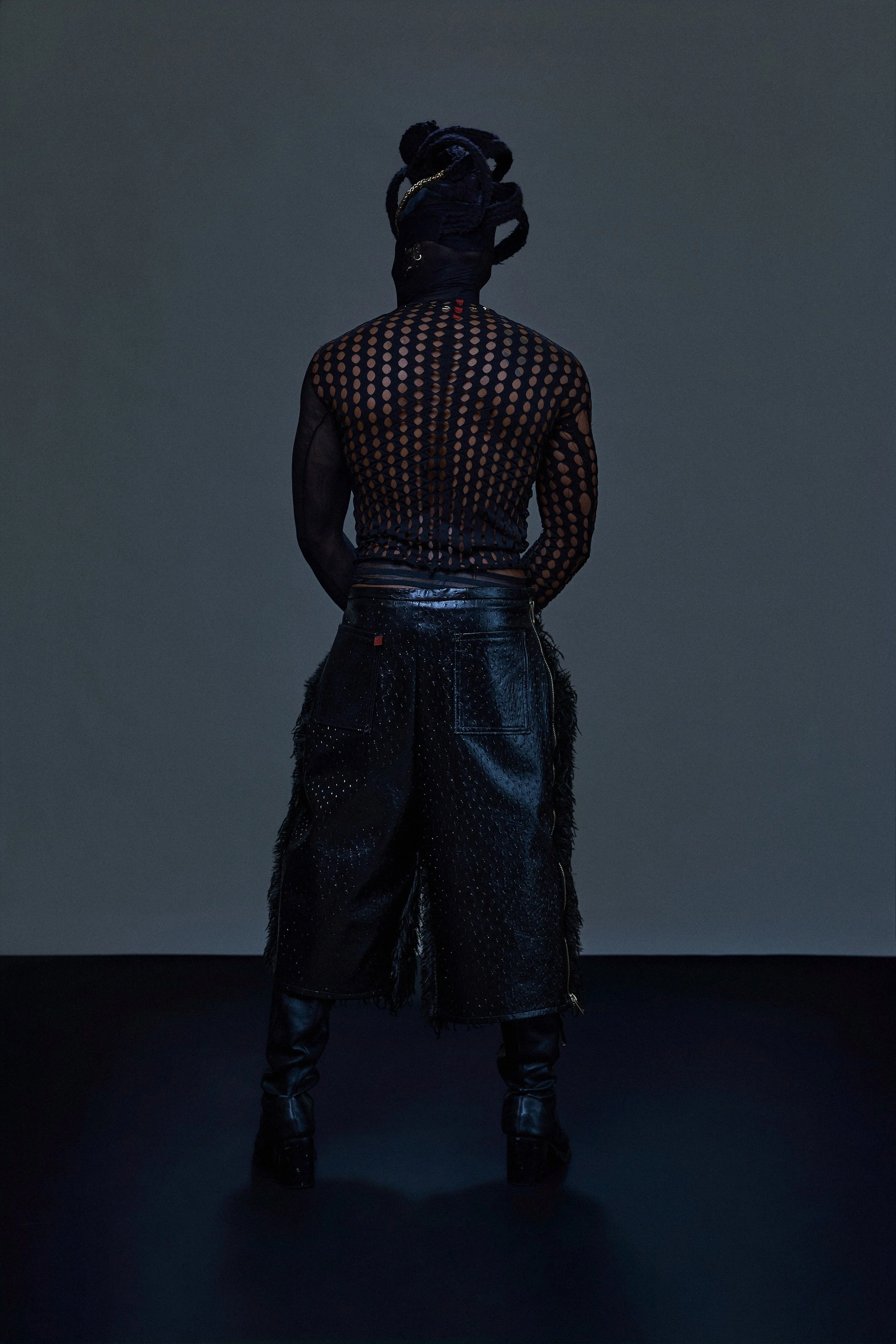 Geoffrey Smith - Person standing with their back to the camera, wearing a black mesh top with circular cutouts, black shiny pants, and black boots, set against a dark, minimal background.