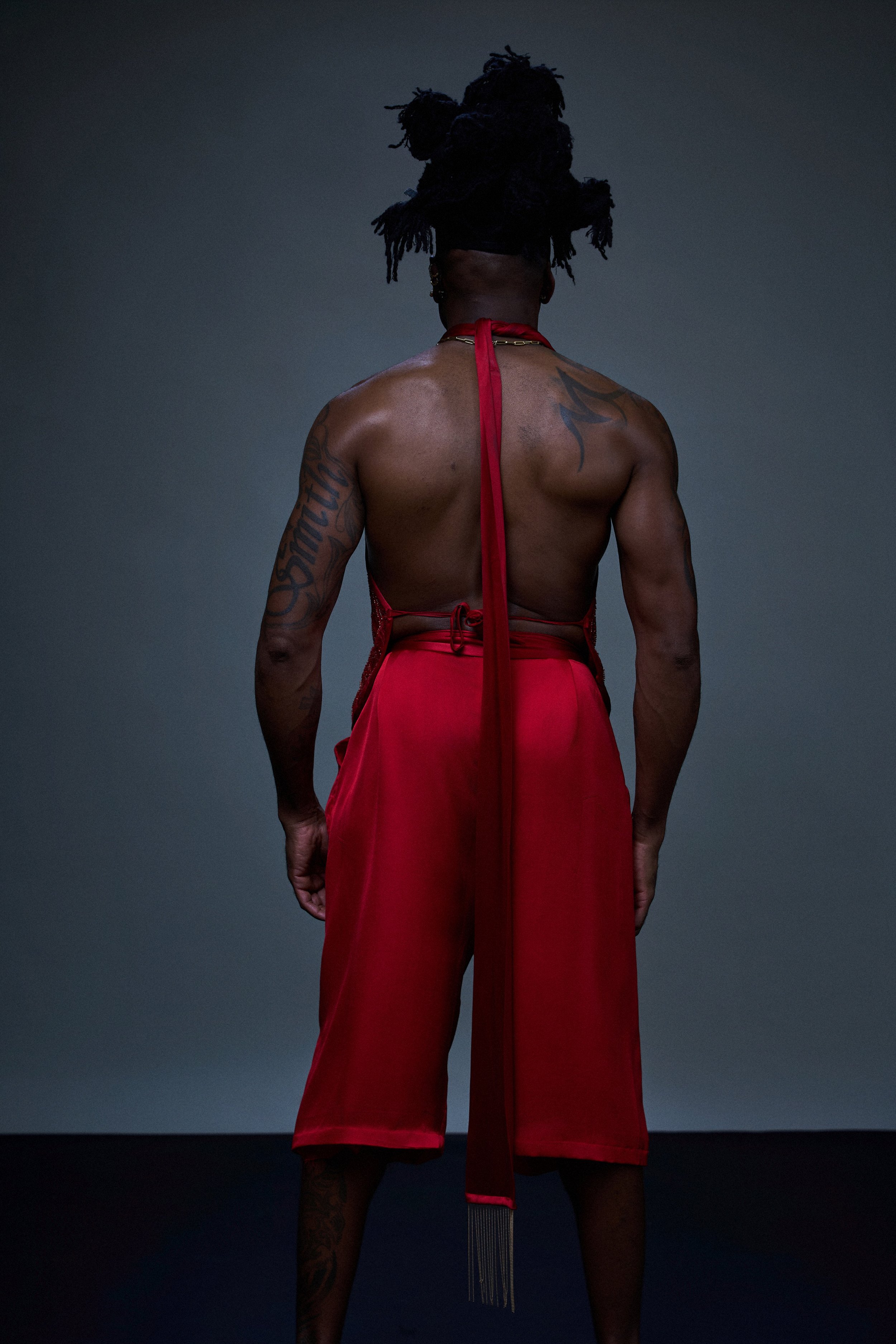 Back view of a shirtless person with tattoos, wearing red pants and a red scarf, standing against a dark background.
