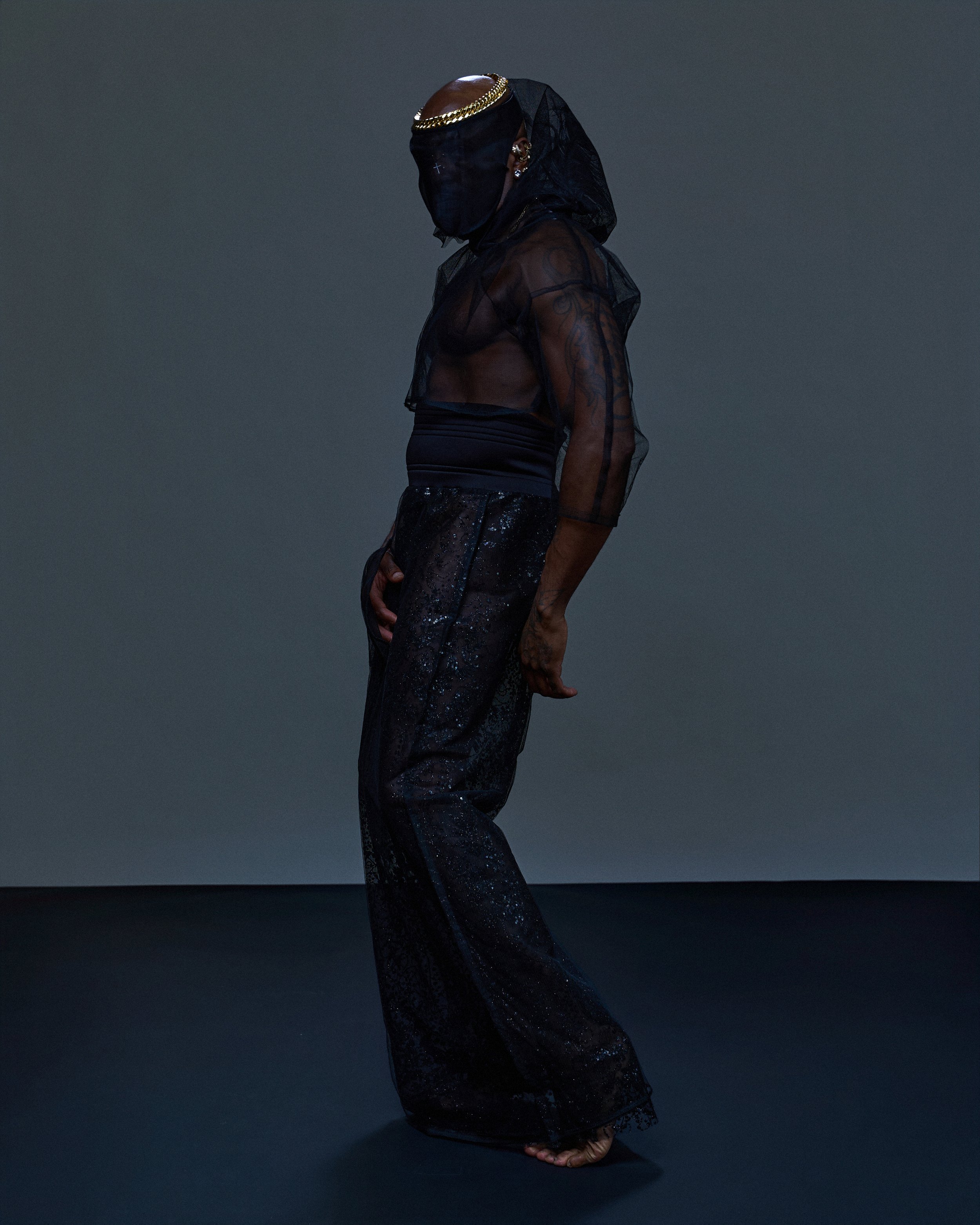 Person wearing a black, sheer, long-sleeve top with tattoos visible on the arm. They have a gold chain headpiece and a black face covering, with earrings and a black, shimmering outfit.