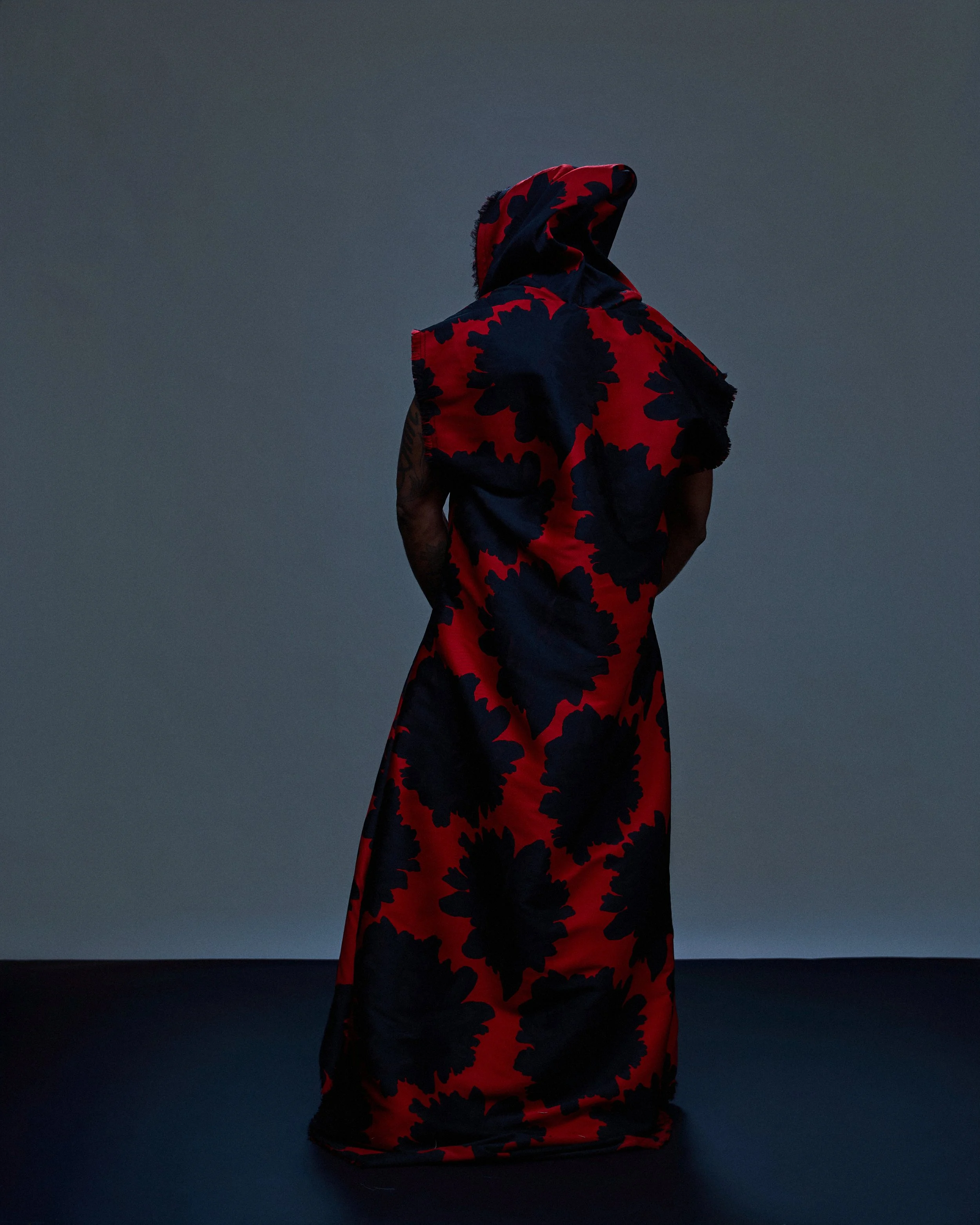 Person wearing a red and black floral patterned robe or shawl, standing against a dark background.