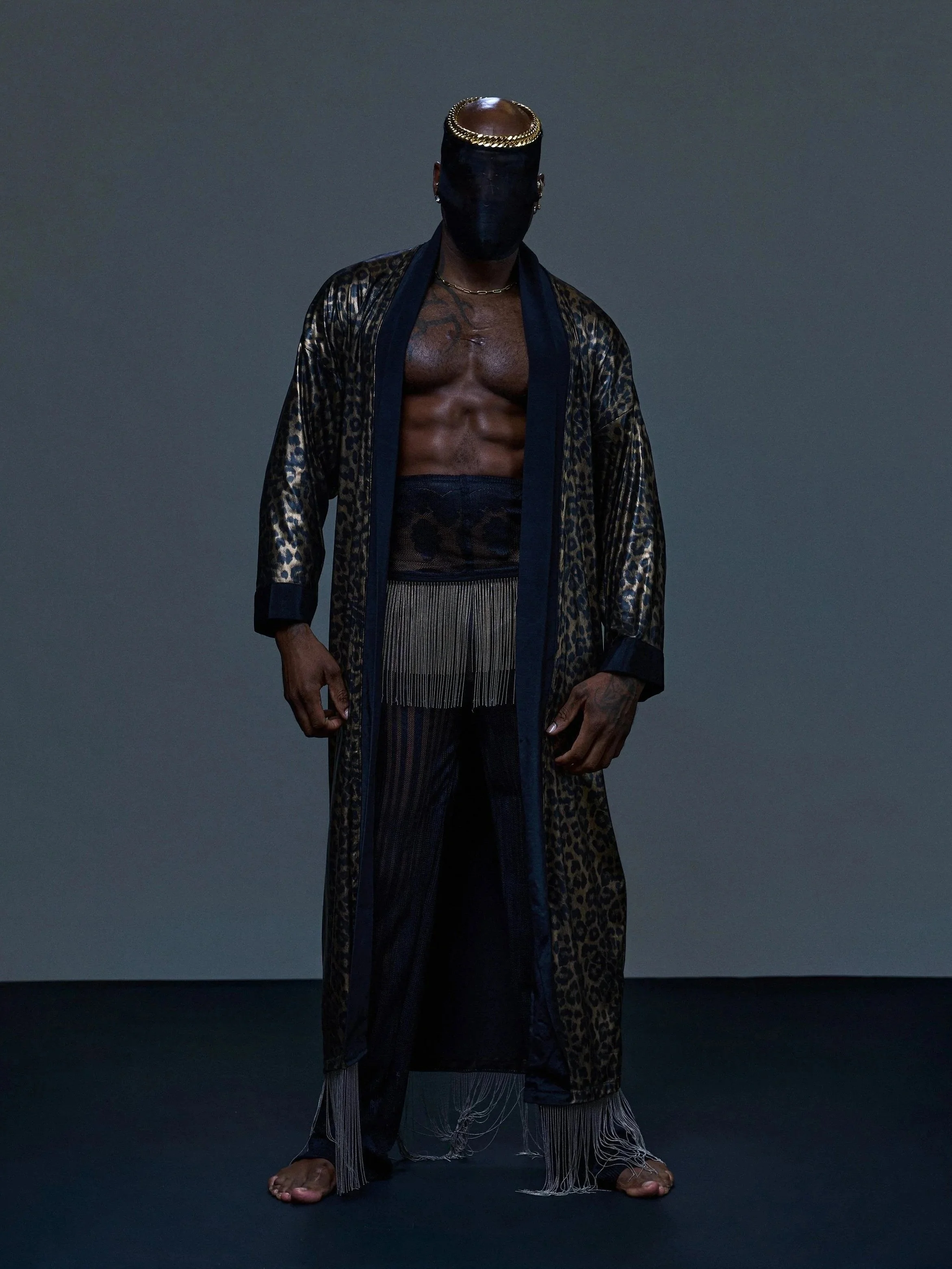 Person with a shaved head wearing a gold-circled black mask, a black and leopard print robe, a gold chain necklace, loose patterned pants, standing on a dark background.