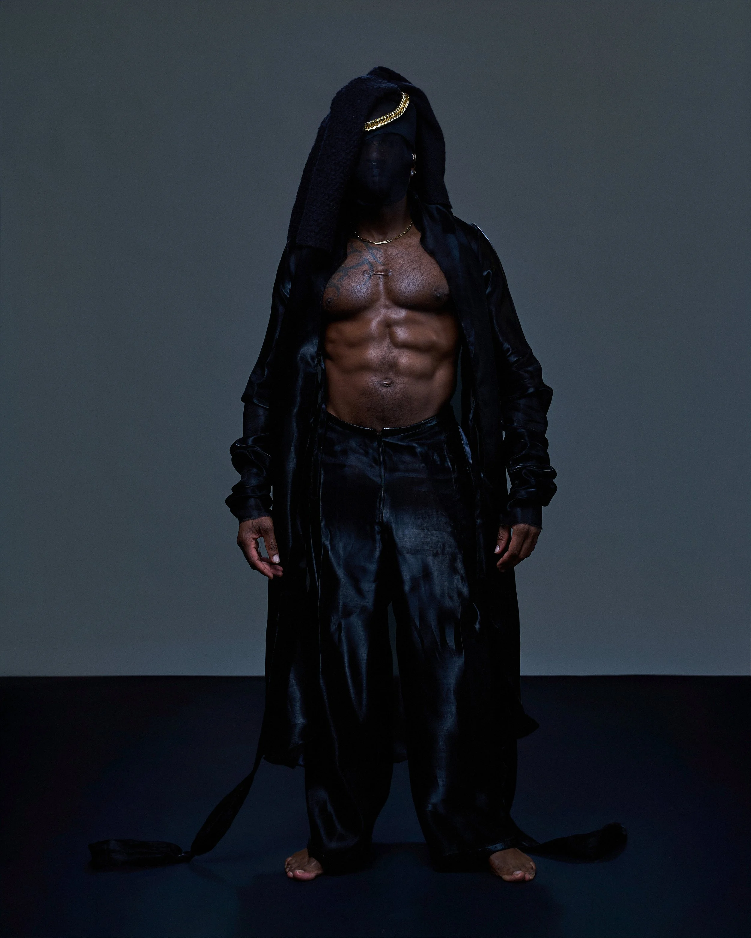 A shirtless man with dark skin is standing against a plain dark background. He is wearing black leather pants, a black jacket open at the front, a gold chain, an earring, and a head towel. His long black hair is covered by the towel, and he is barefo
